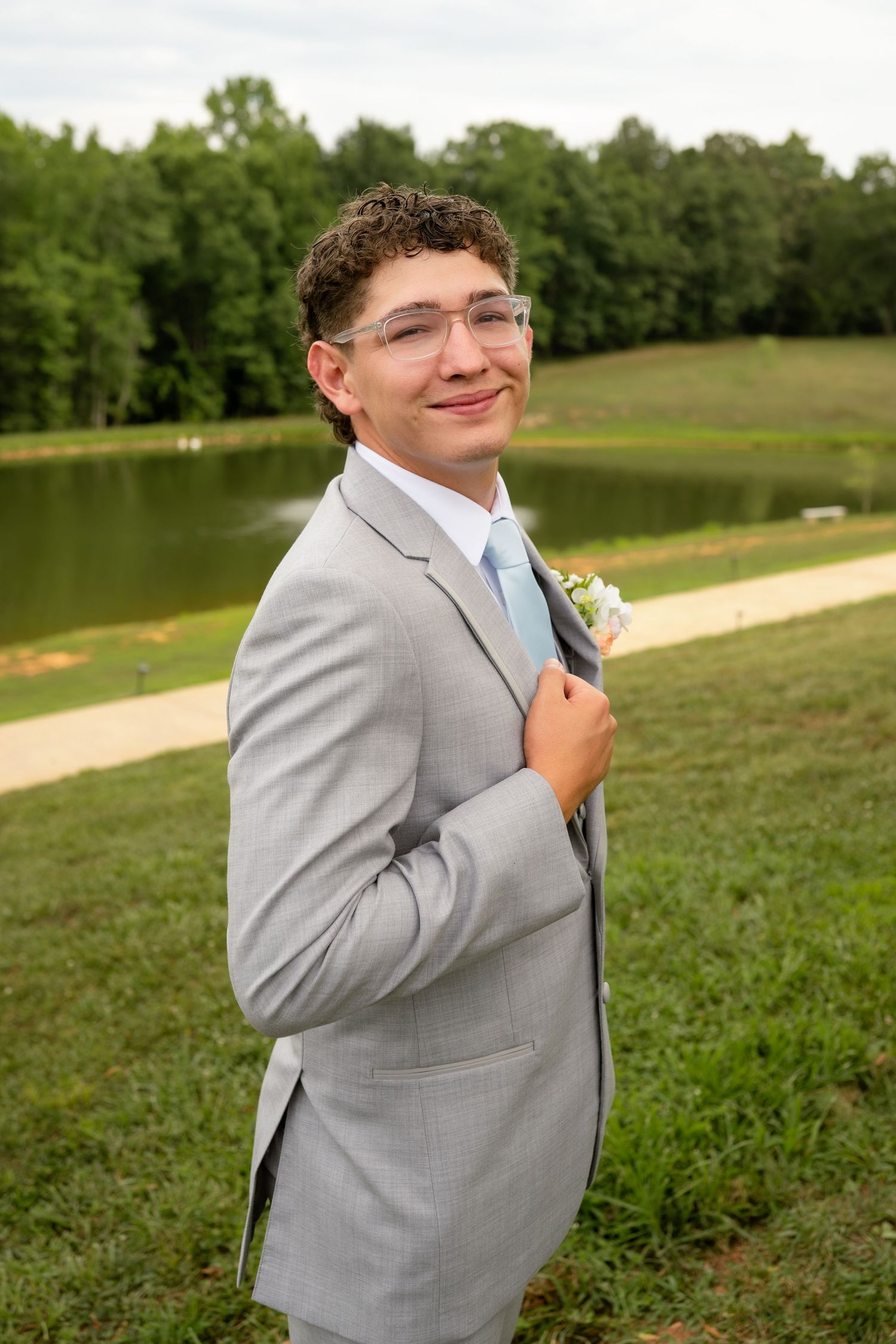 Smiling young groomsman in grey suit adjusts boutonniere while posing outdoors near a serene pond at wedding.
