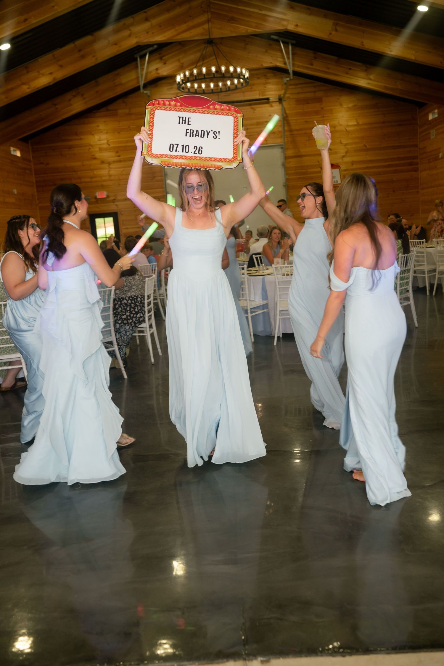 Bridesmaids in light blue gowns celebrate at wedding reception holding glow sticks and a personalized sign.