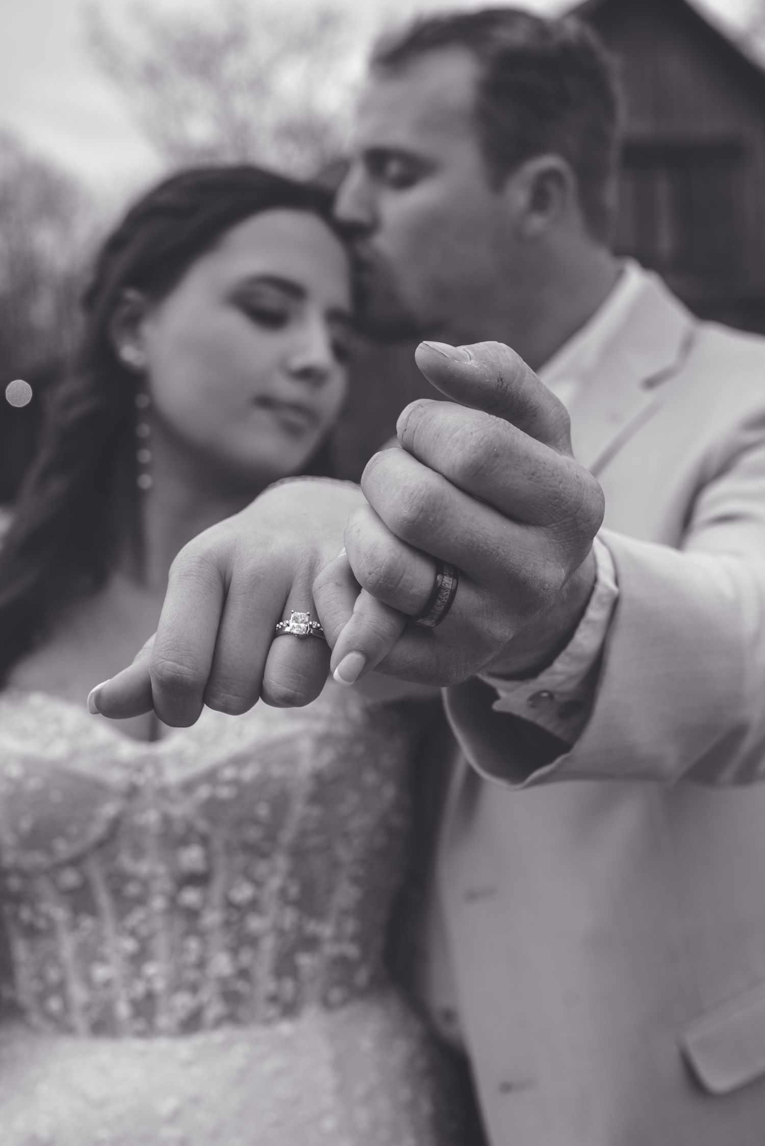 Black and white wedding photo of bride and groom showing off their rings, groom kissing bride on the head.