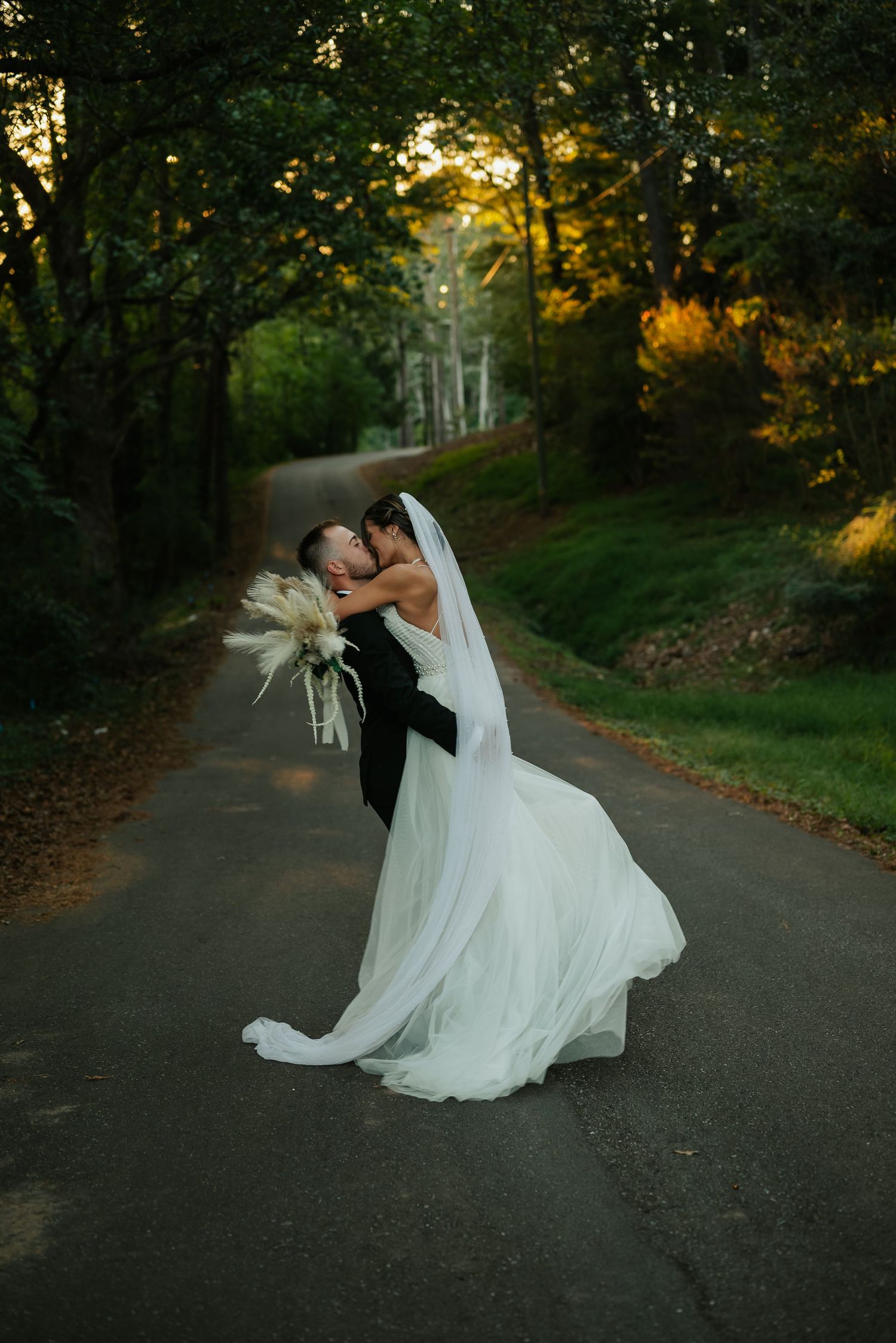 A couple in wedding attire share an intimate dip pose on a tree-lined road at sunset. Southern pines, NC  Wedding Photographer