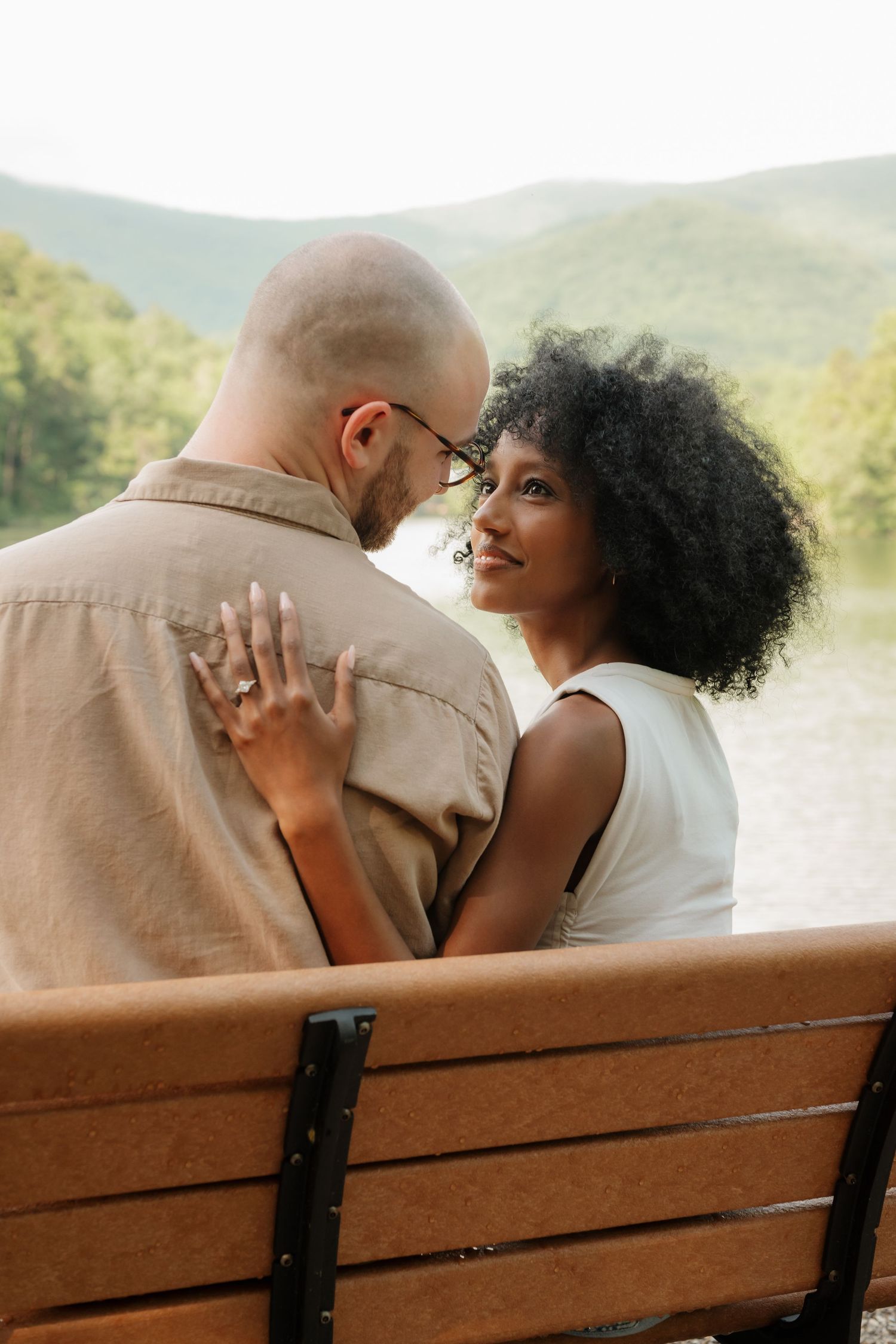 A romantic couple sits close together and shares intimate moments on a wooden park bench with mountains in the background. in North Carolina
