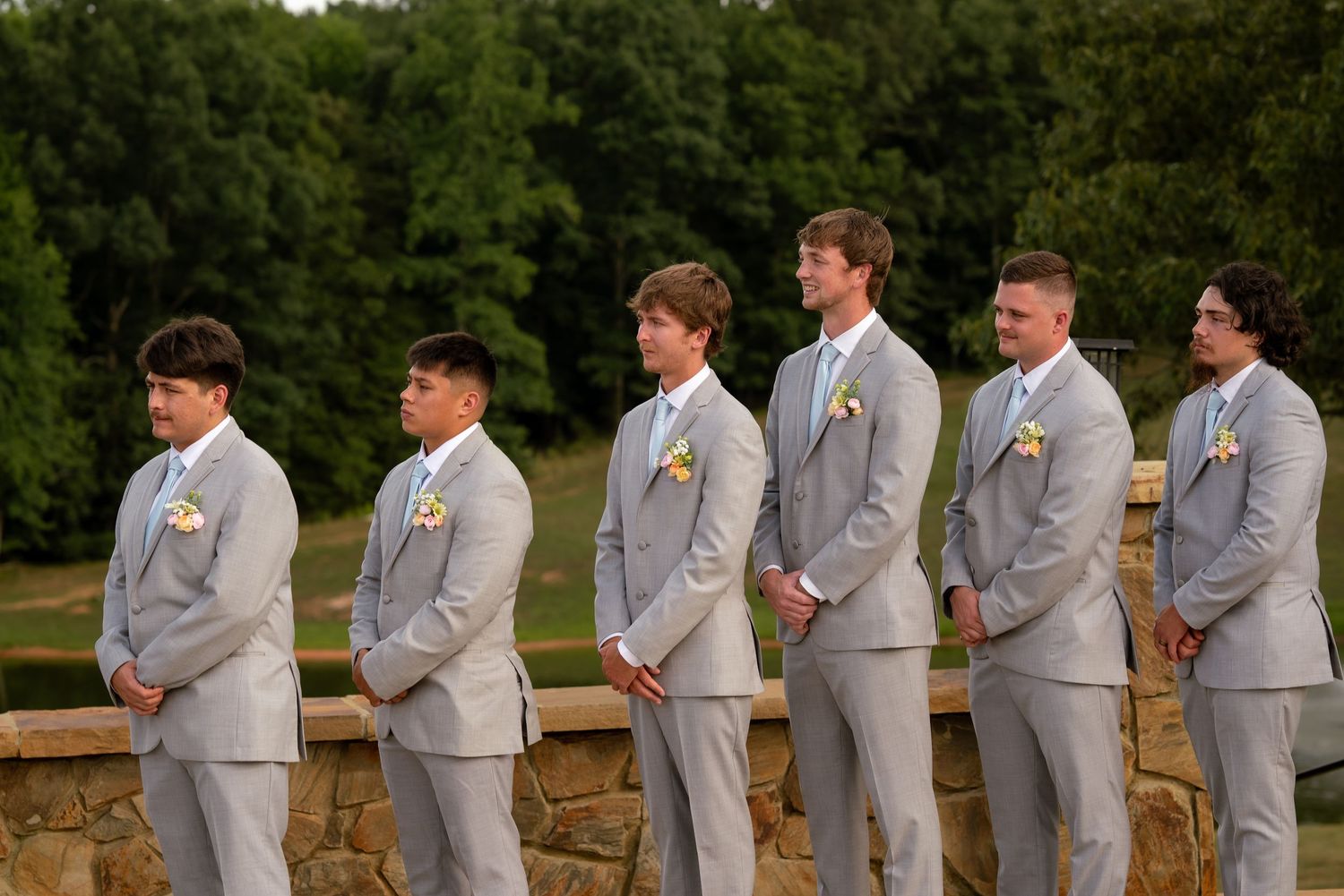 Six groomsmen in matching light gray suits with boutonnieres stand in a line outdoors at a wedding venue.