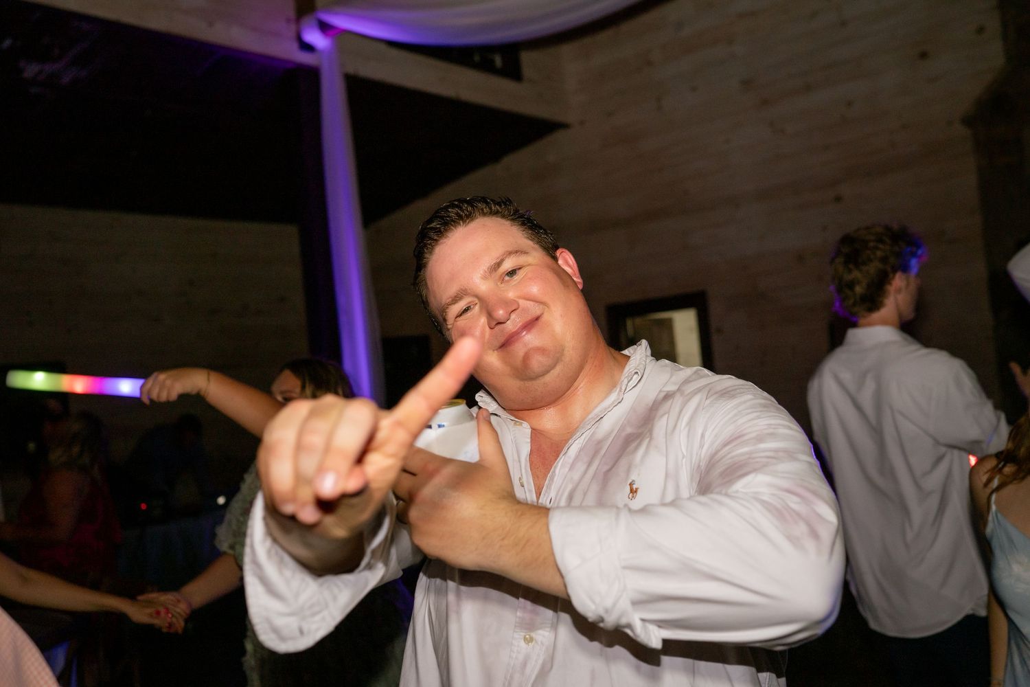 Man in white shirt dancing and pointing at camera at a lively indoor party with colorful lights.