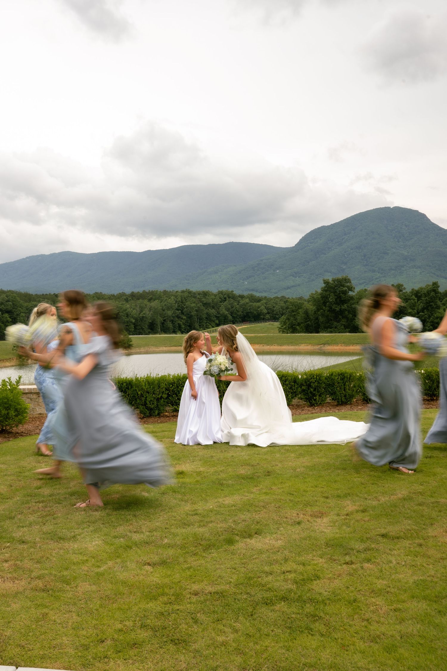 Bride kisses flower girl while bridesmaids in blue-gray dresses run past with mountain backdrop.