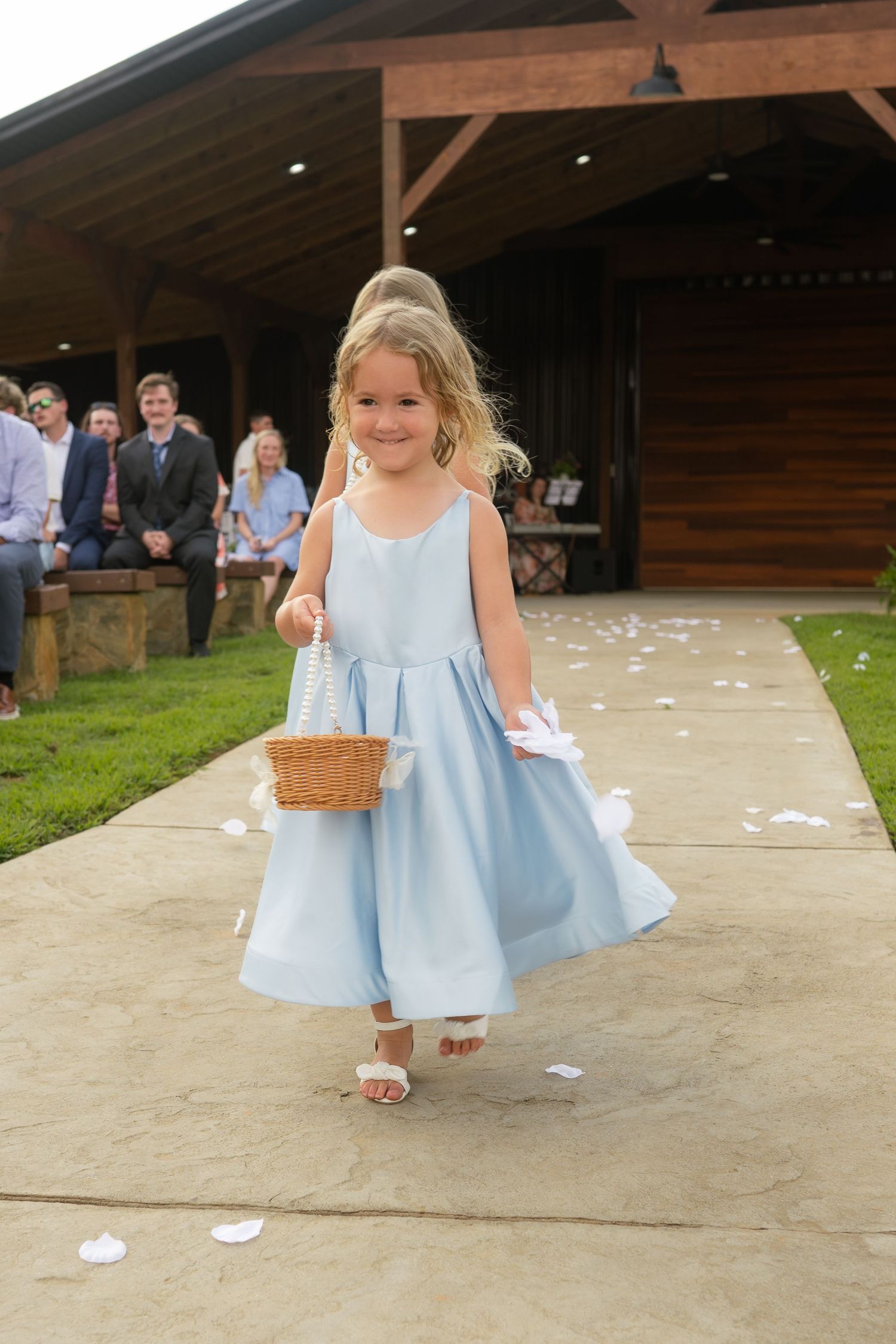 Young flower girl in light blue dress walks down wedding aisle scattering white petals from wicker basket.