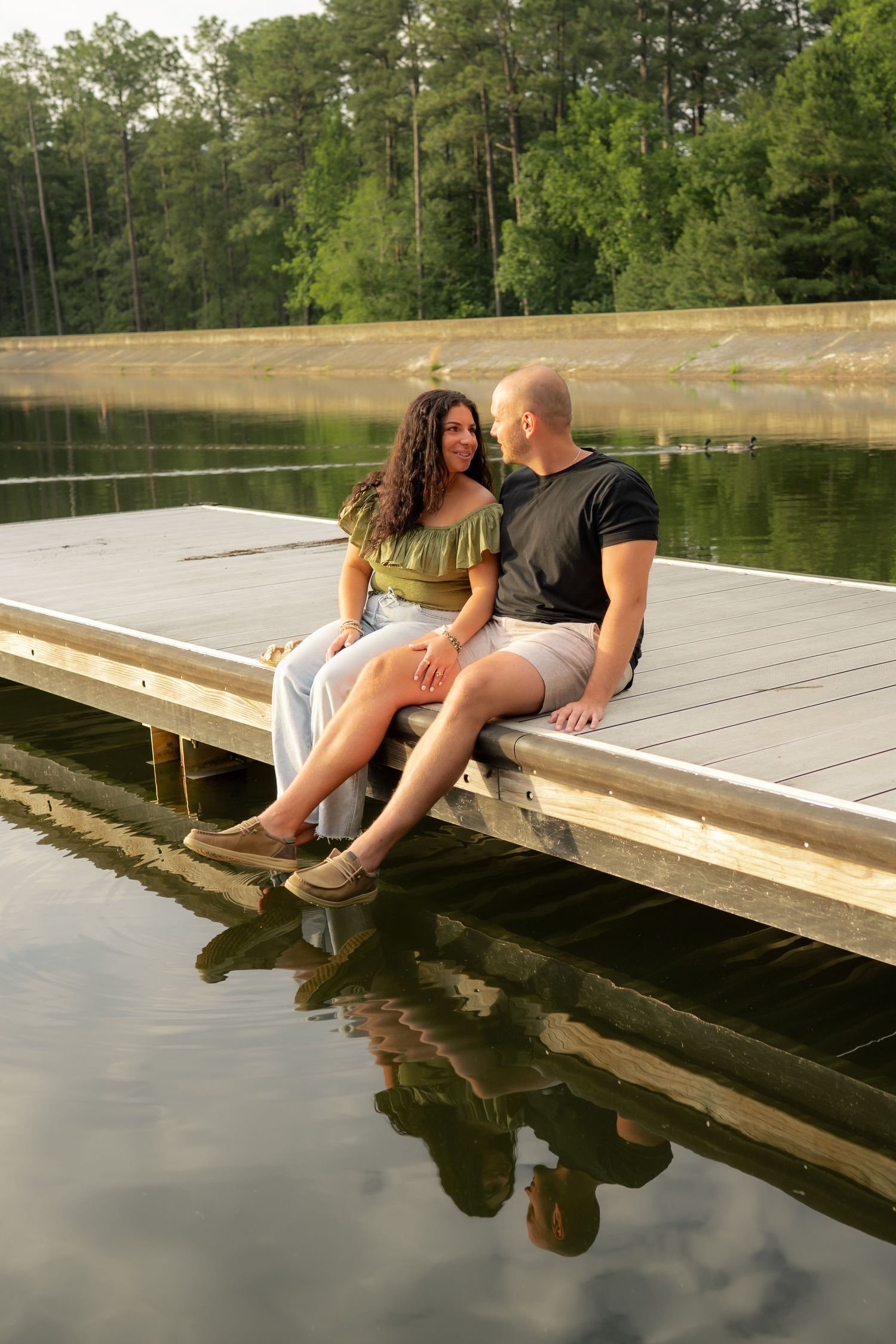 Couple sitting together on a wooden dock by a calm lake, surrounded by lush green trees, sharing a romantic moment.