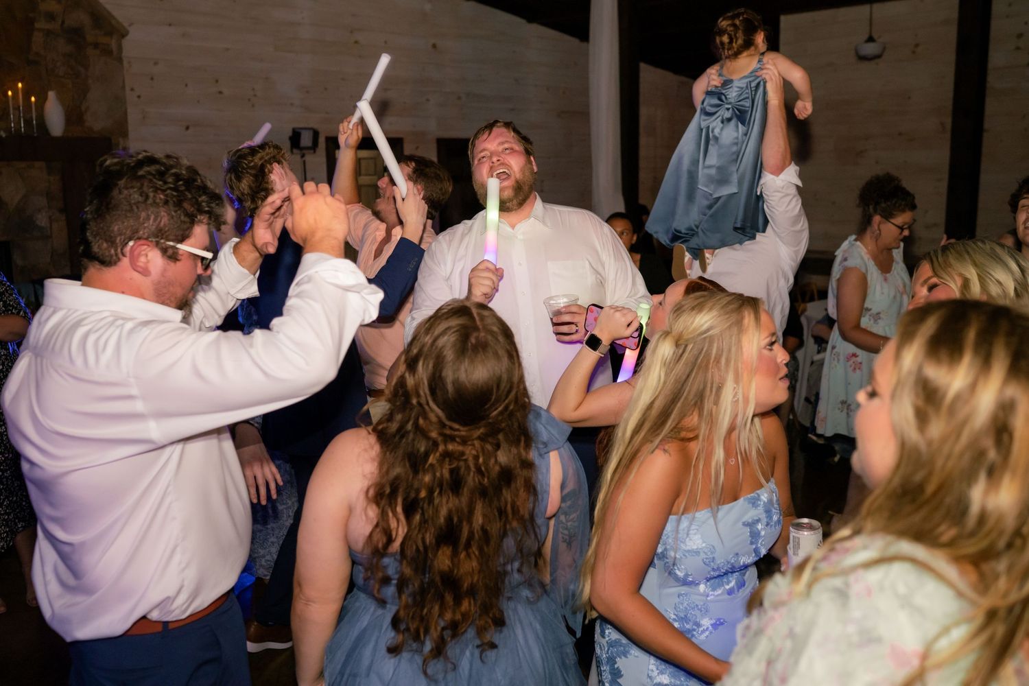 Energetic wedding guests dancing with glow sticks at lively indoor reception party.