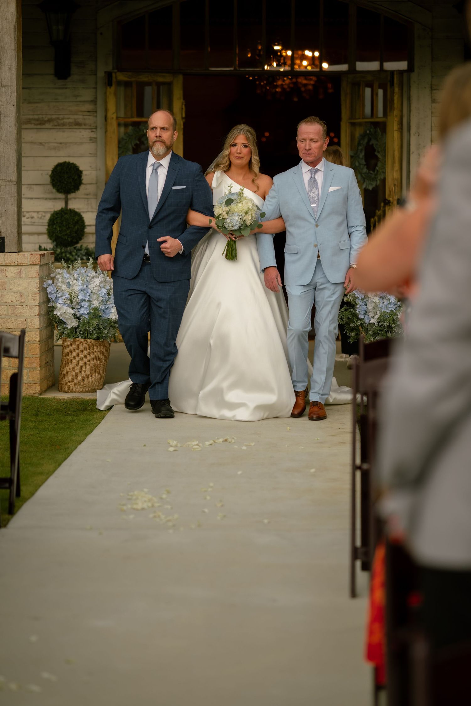 Bride in white ball gown walking down aisle escorted by two men in navy and light blue suits at outdoor ceremony.