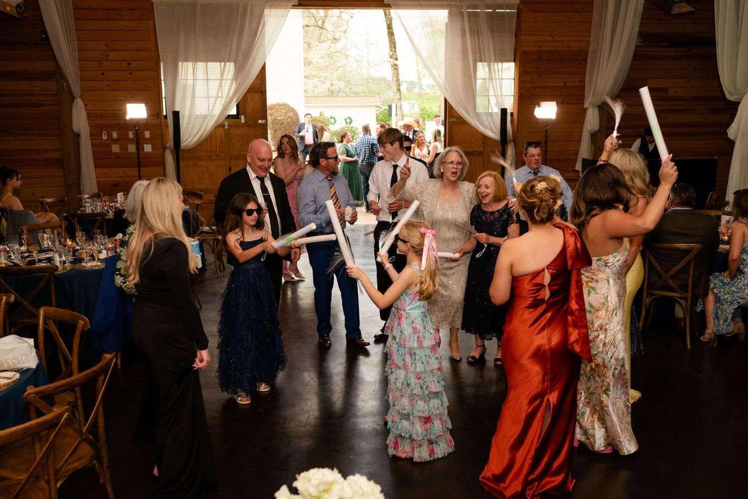 Wedding reception guests dancing and celebrating with light-up batons inside a rustic barn venue with white draping.