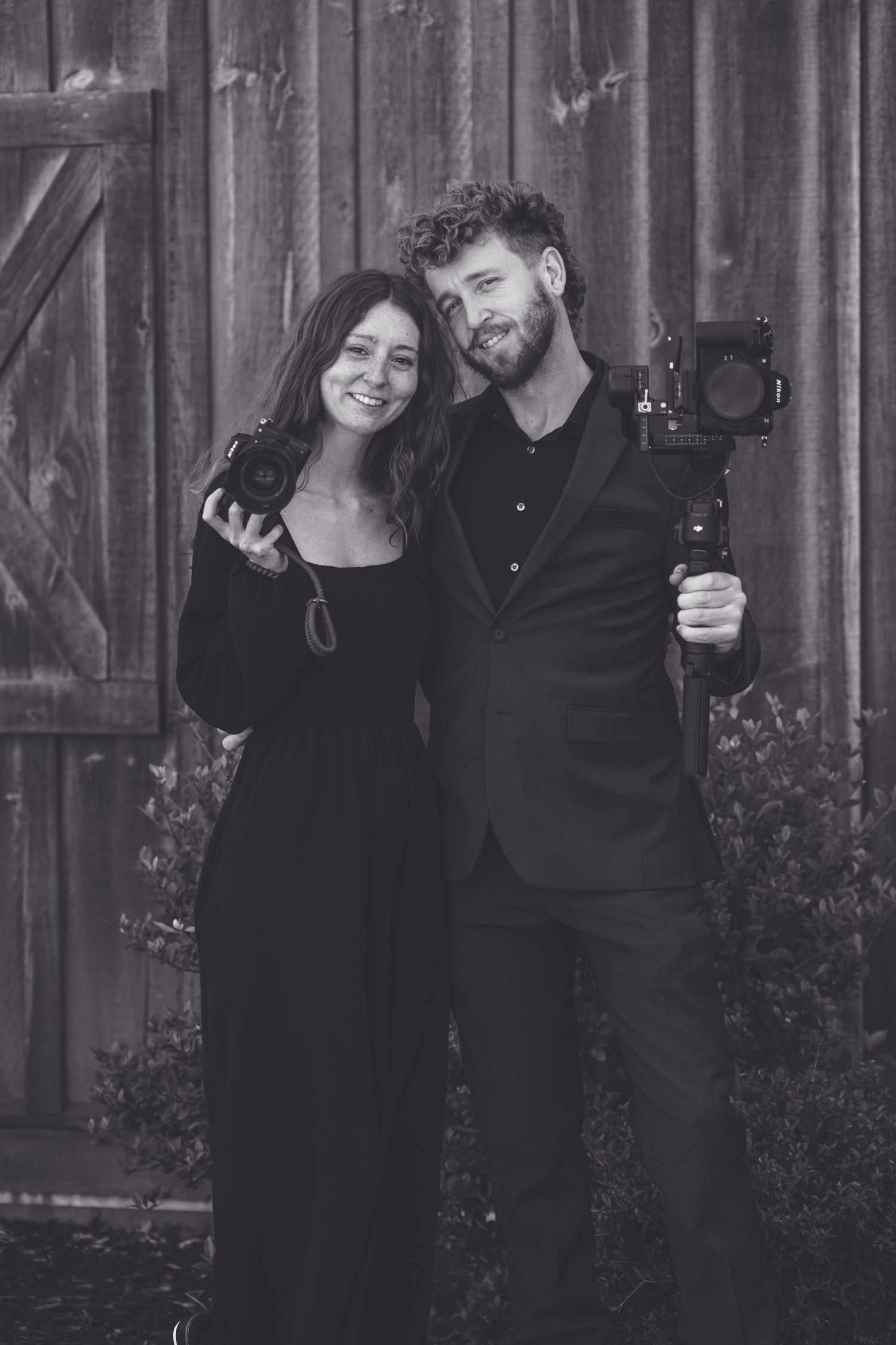 Black and white photo of two photographers standing together with their cameras against a rustic wooden wall.