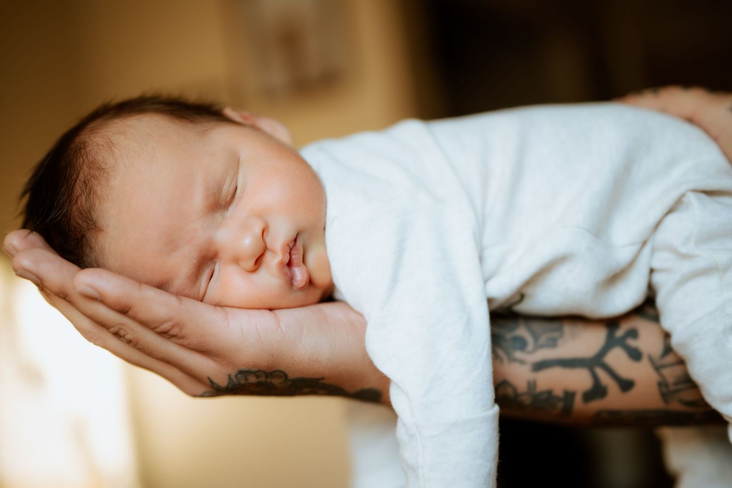 Sleeping newborn in white onesie resting peacefully on tattooed arms.