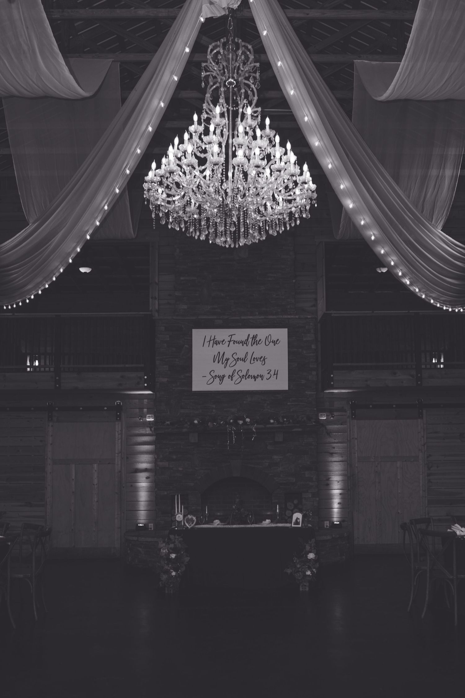 Black and white photo of elegant barn wedding reception with grand chandelier, draped fabric, and Song of Solomon sign.