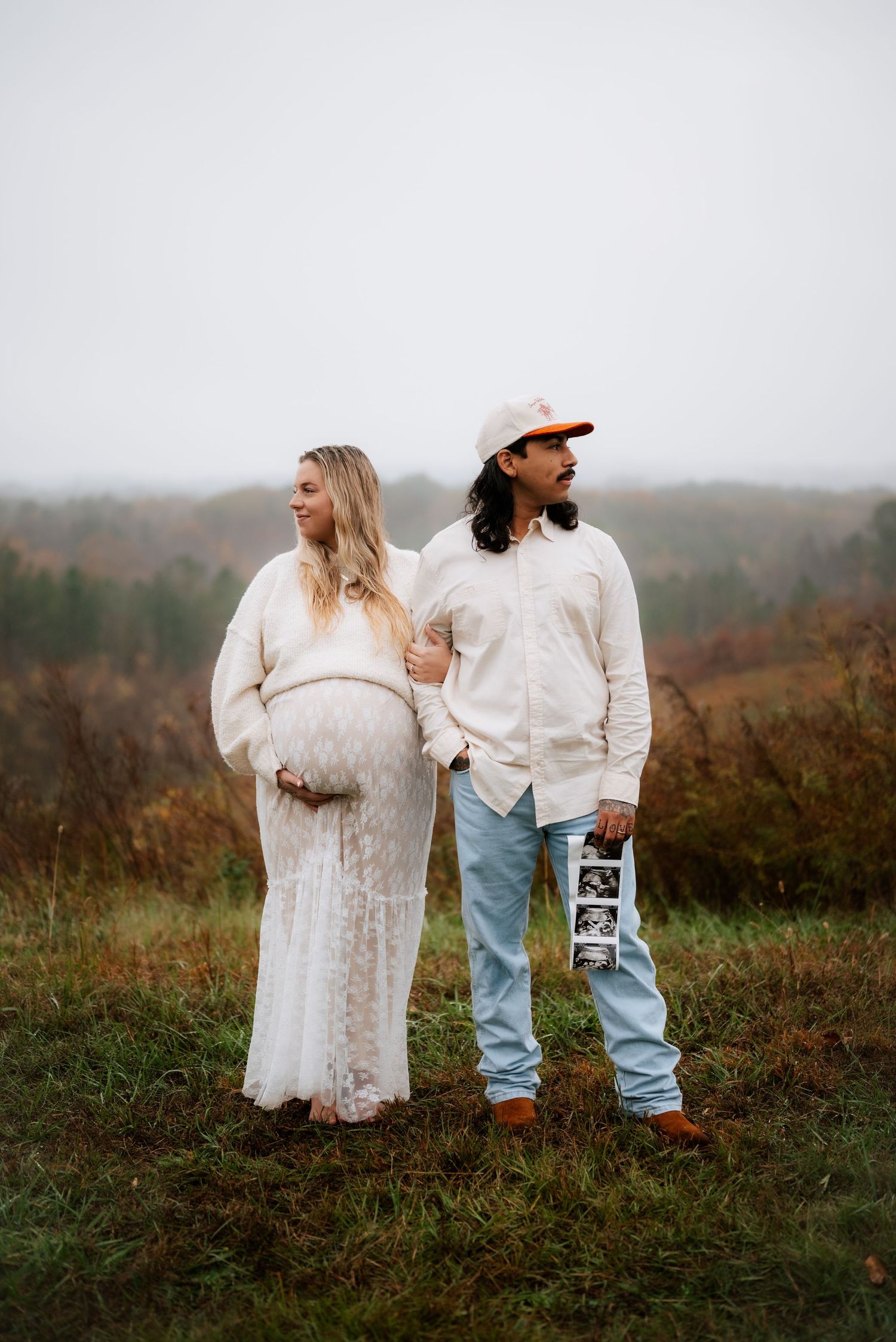 Expectant couple in matching white outfits standing in autumn field.