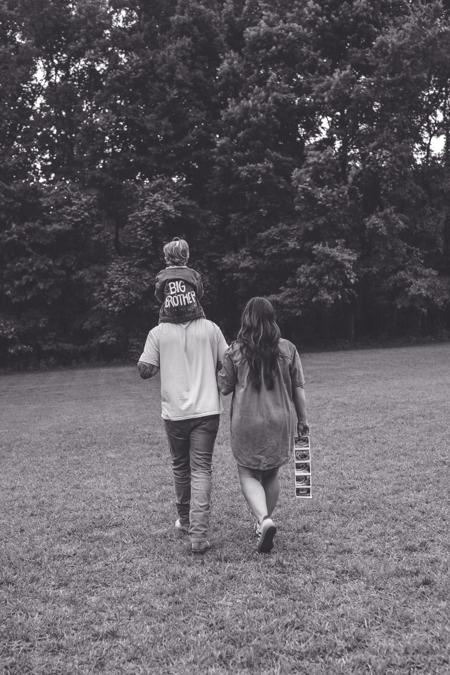 Black and white photo of a family walking in a park, with a child on dad's shoulders wearing a Big Brother shirt.