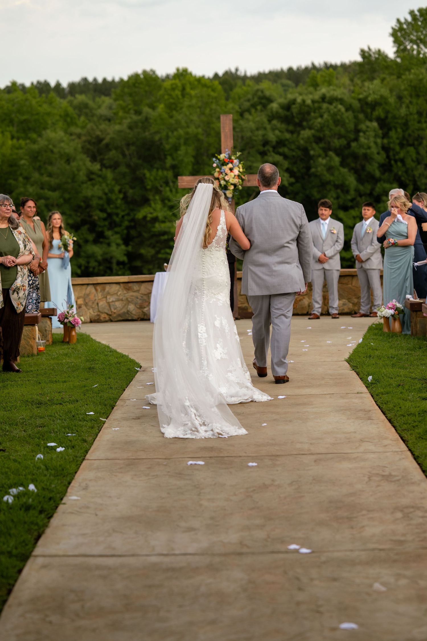 Bride walks down outdoor wedding aisle with veil trailing, guests watching, wooden cross at altar amid lush greenery.