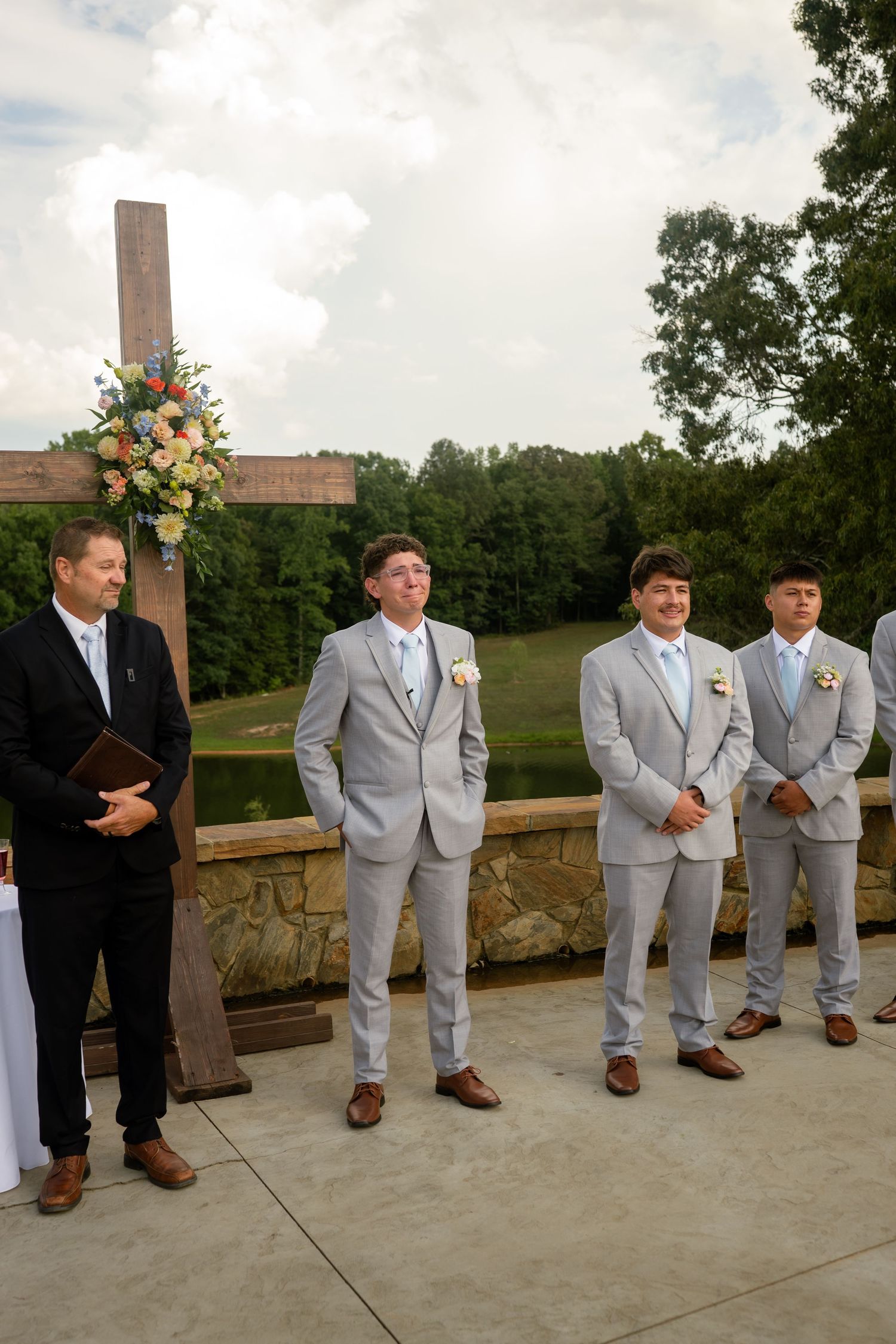 Groomsmen in light gray suits stand at outdoor wedding altar beside wooden cross with colorful flowers.