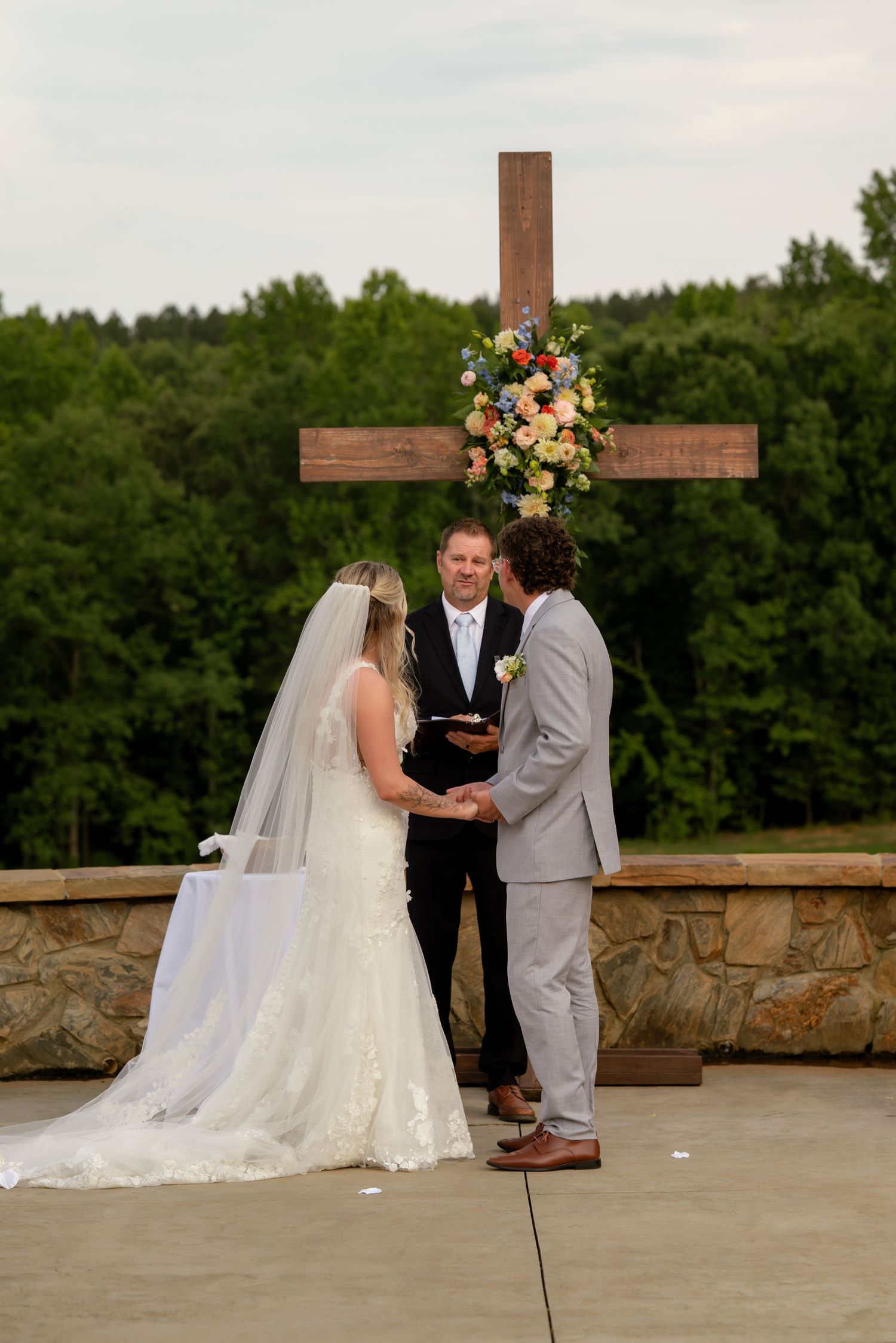 Outdoor wedding ceremony at a stone altar with a wooden cross adorned with colorful florals and lush greenery.