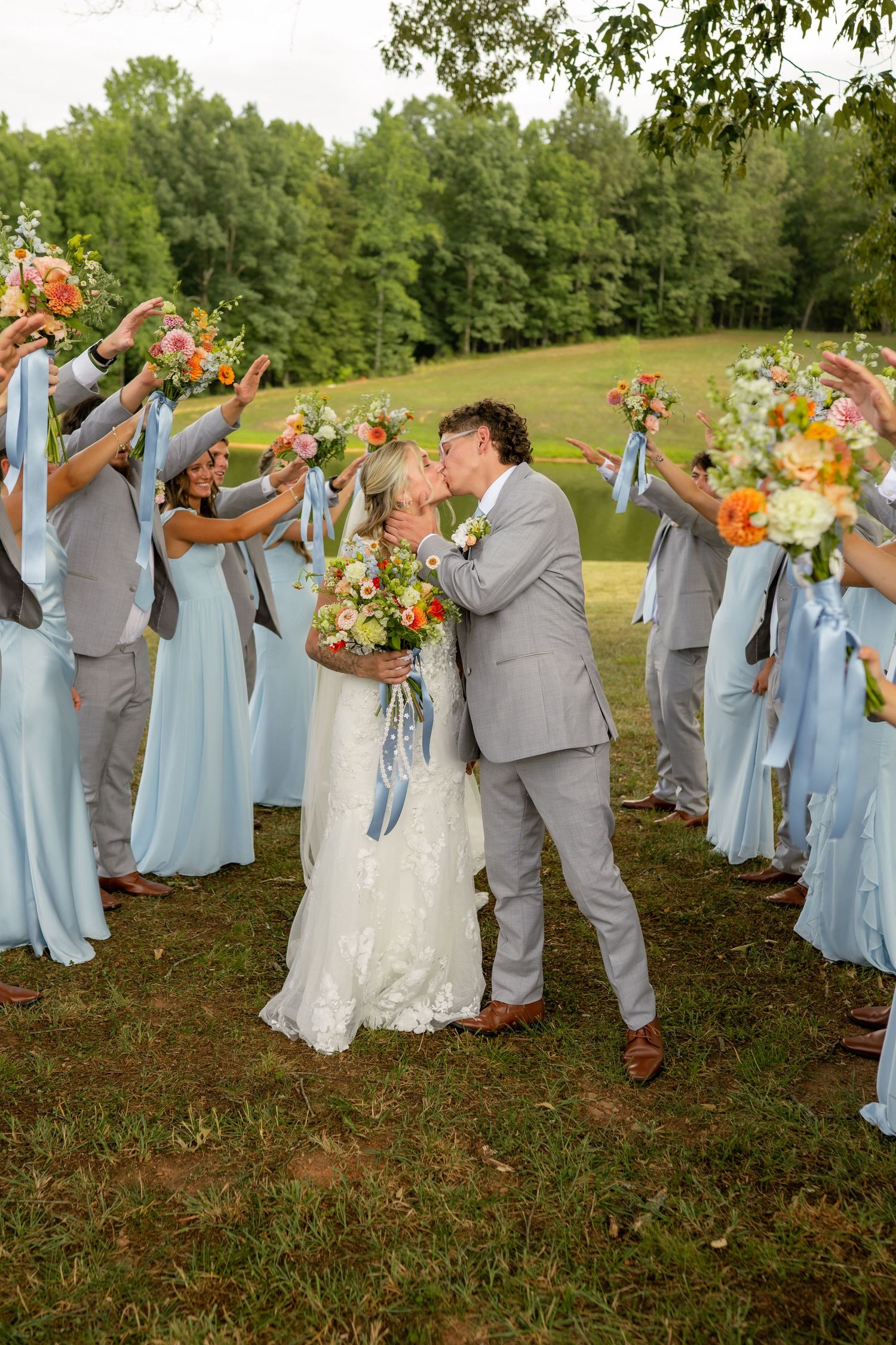 Bride and groom kissing while wedding party in blue dresses and gray suits raise colorful bouquets overhead.