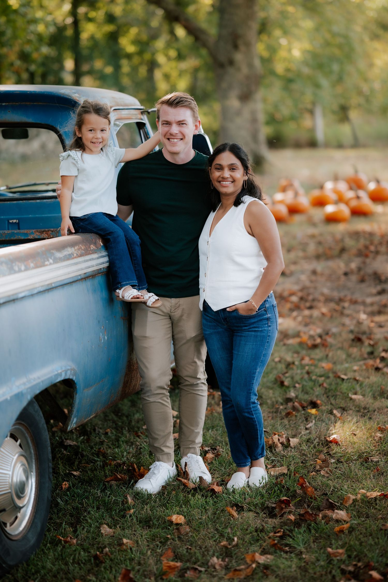 Family poses beside classic blue pickup truck with pumpkins in autumn setting.