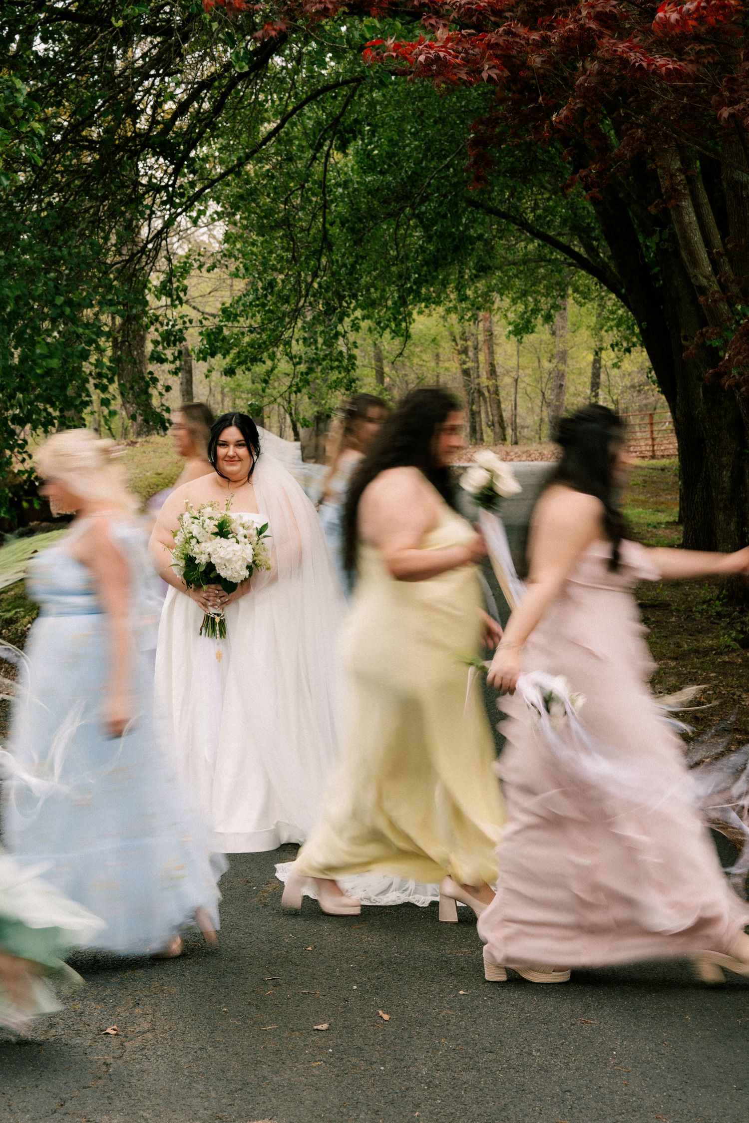 Motion-blurred wedding party in long dresses walks together on a tree-lined garden path as flowers bloom overhead. North Carolina
