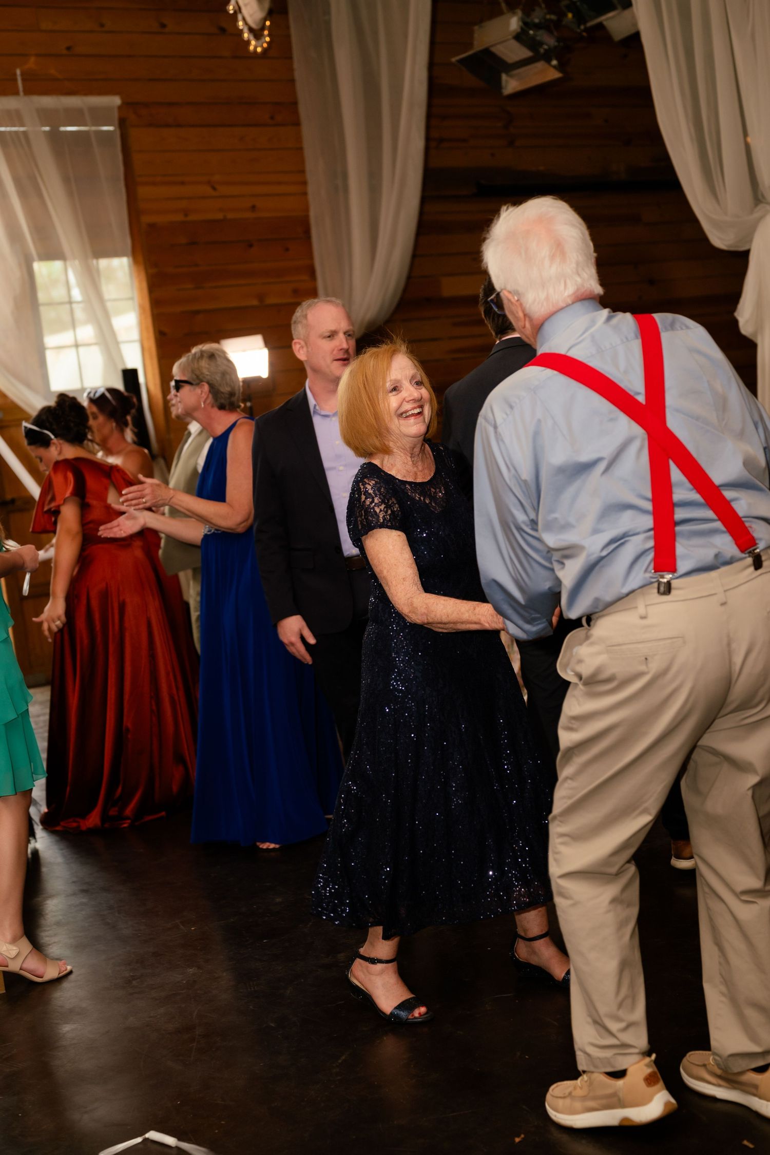 Elderly couple dancing joyfully at a wedding reception, man wearing red suspenders, in a rustic wood-paneled venue.