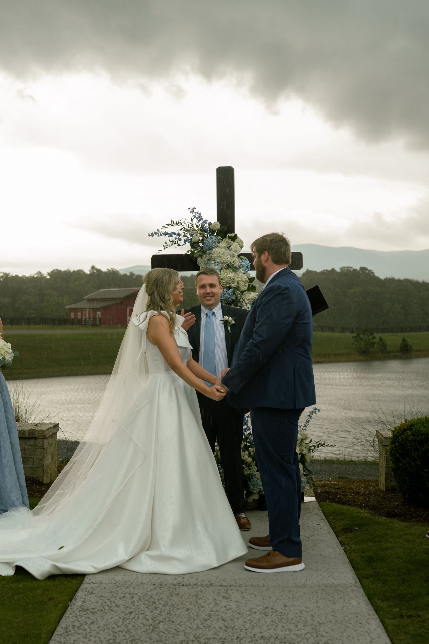 A bride and groom exchange vows before a flower-decorated cross with a scenic mountain lake backdrop on their wedding day.