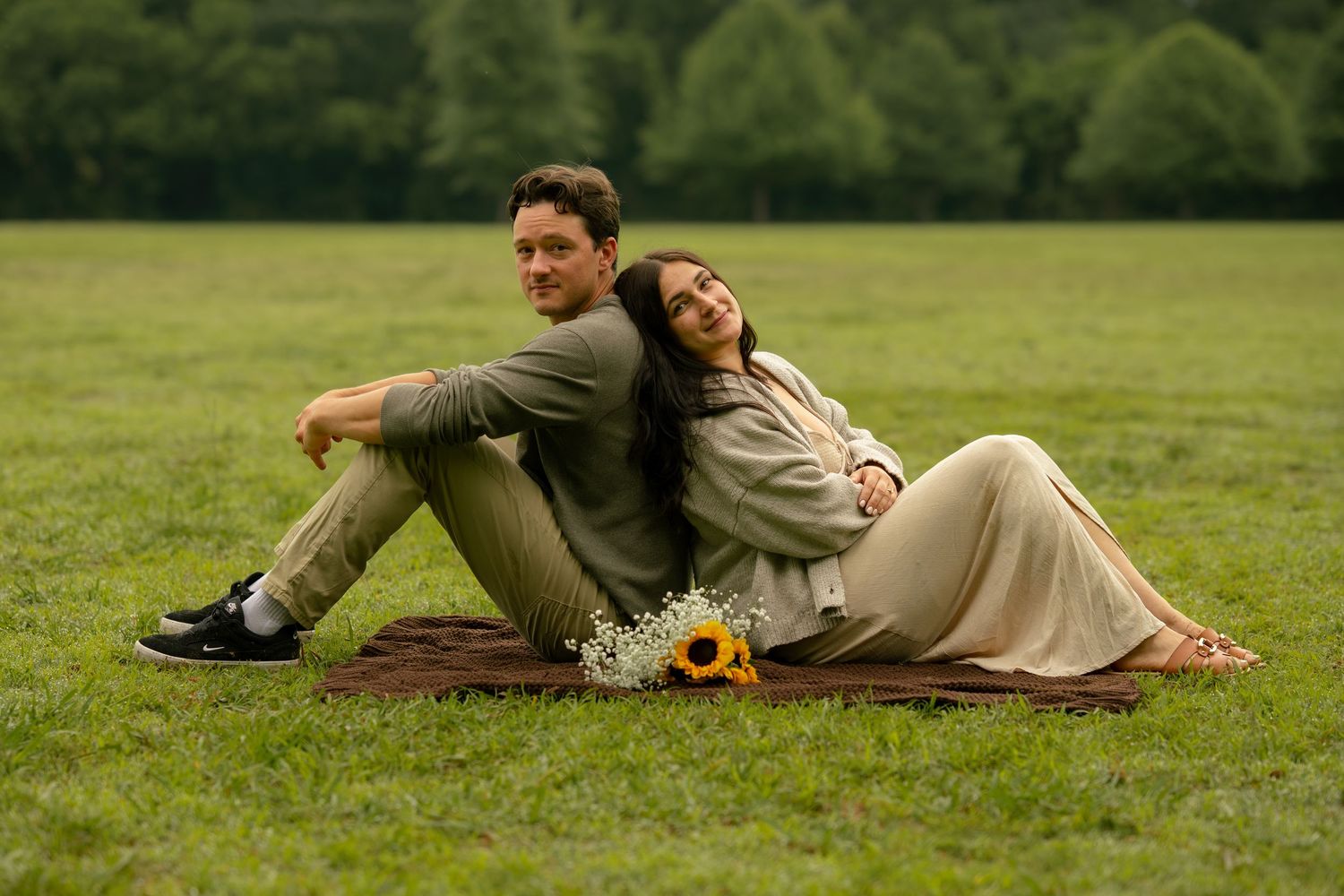 Couple sitting back-to-back on a blanket in a green field with sunflowers, sharing a romantic outdoor moment.