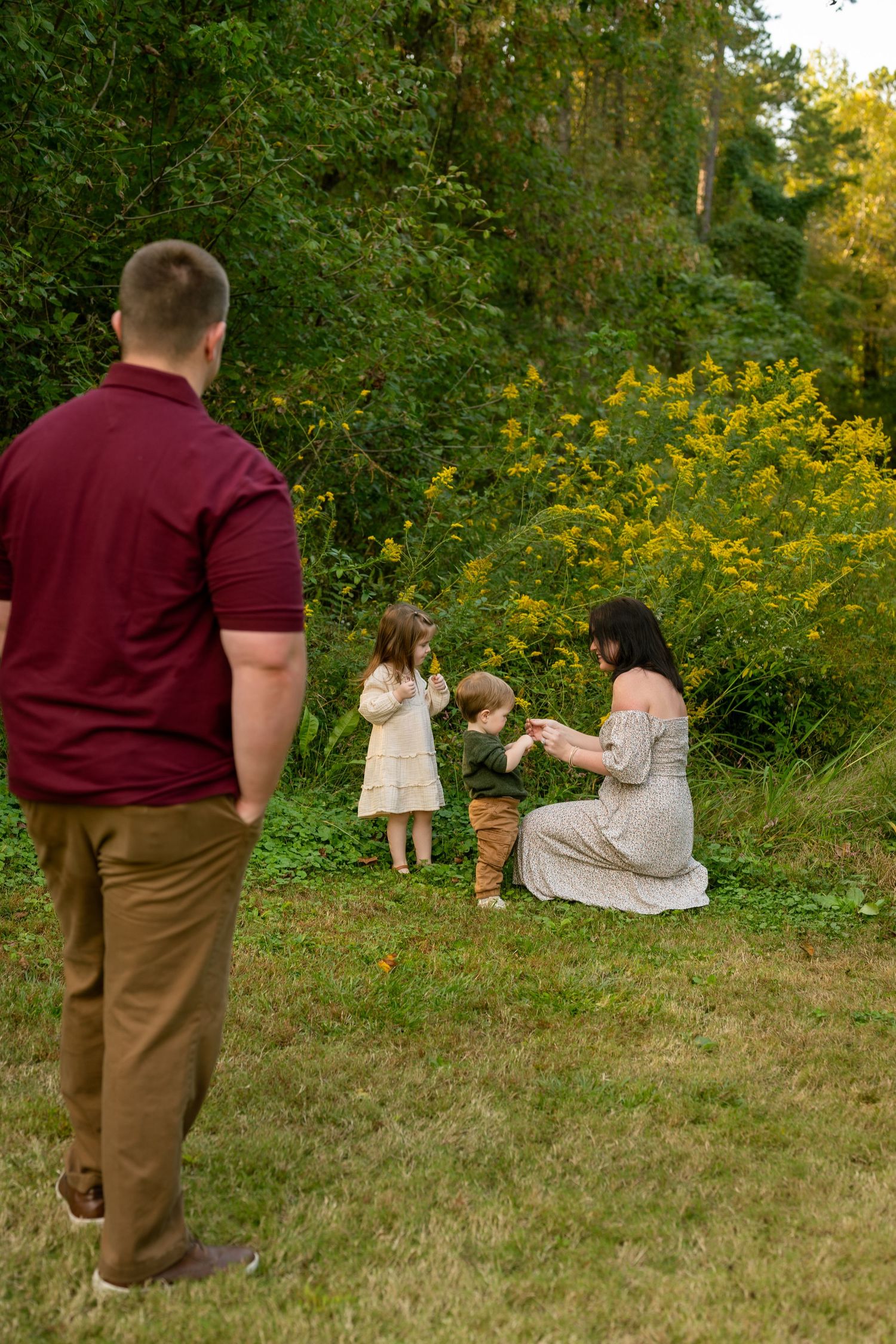 Family outdoors, father watching mother kneel with two young children near yellow wildflowers and green trees at golden hour.