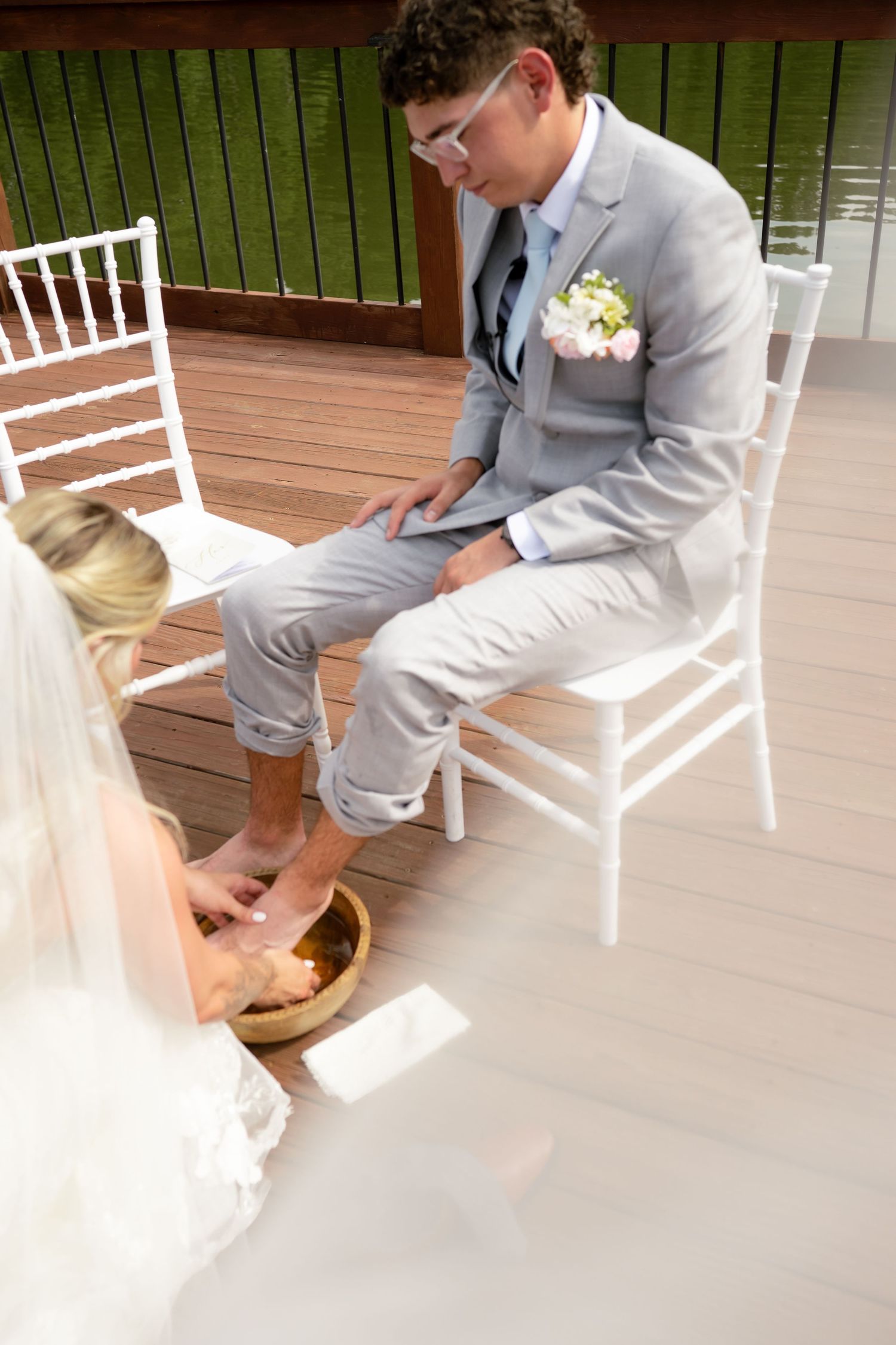 Groom in gray suit has feet washed by bride in white dress during outdoor wedding foot washing ceremony.