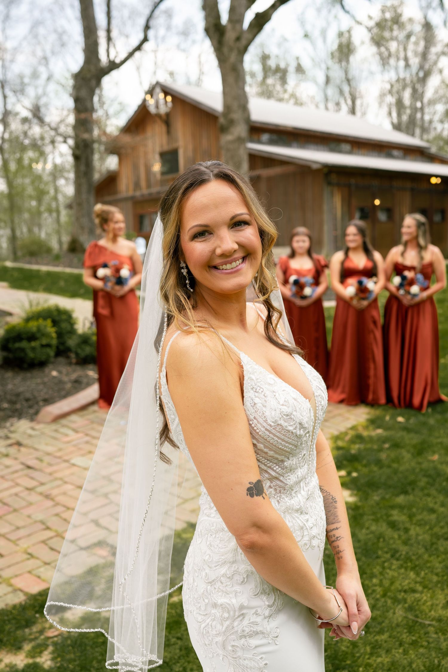 Smiling bride in white lace gown and veil poses outdoors with bridesmaids in rust-red dresses before a rustic barn.