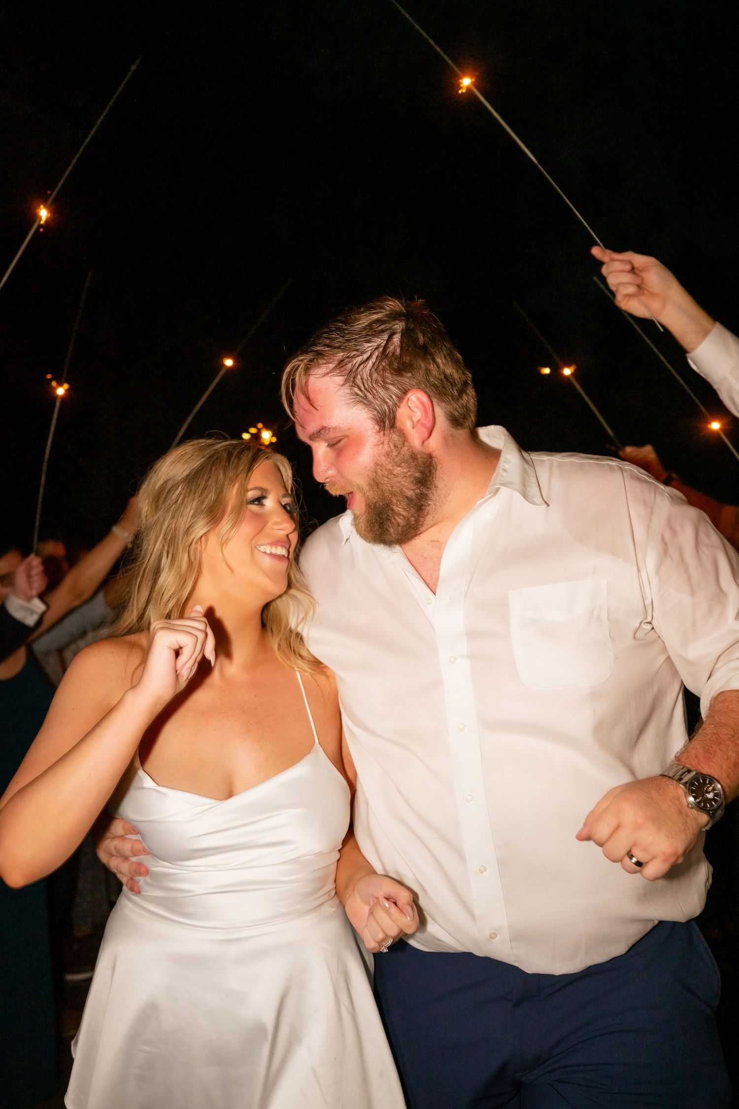 Bride and groom walk through a sparkler exit tunnel at night, smiling joyfully during their wedding reception.