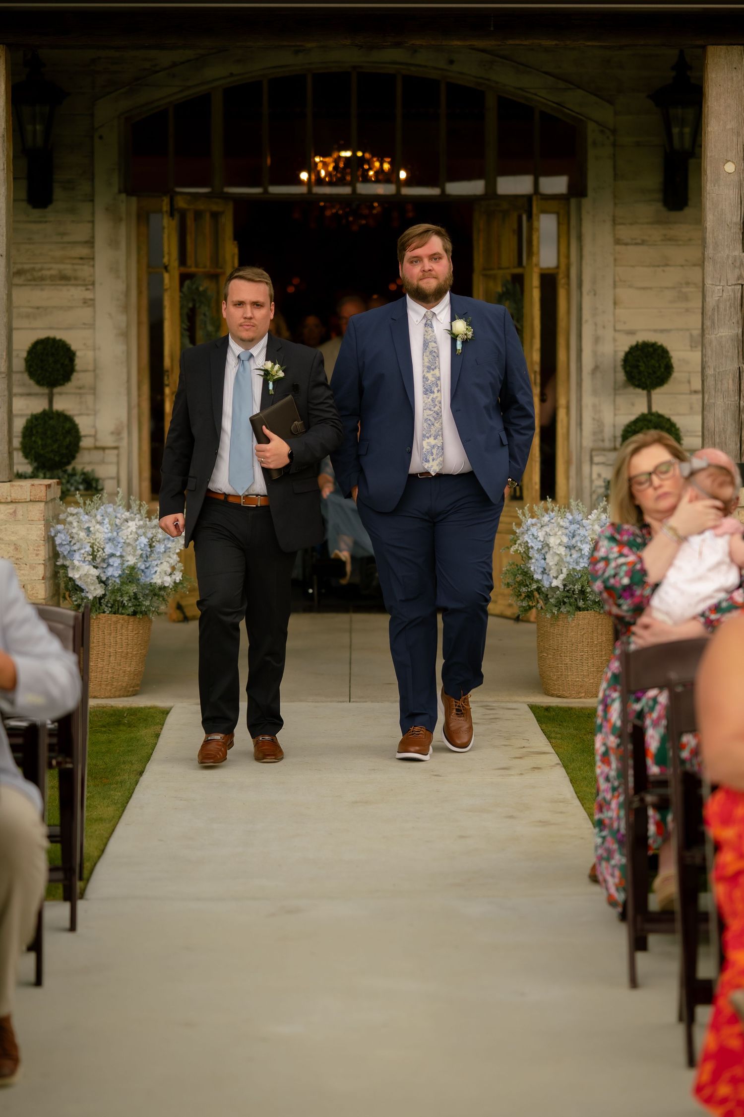 Two groomsmen in navy suits walking down outdoor wedding ceremony aisle.