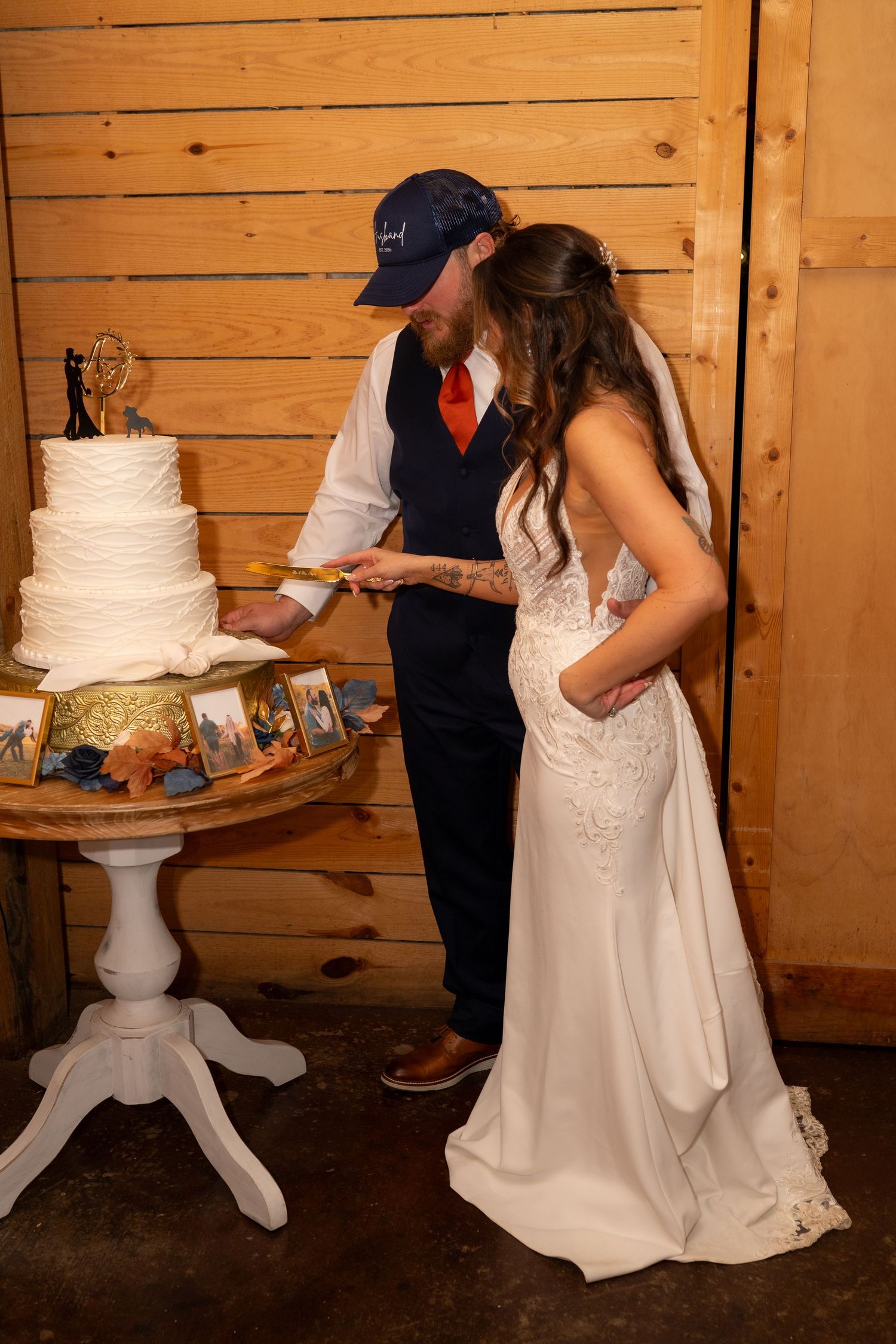 Bride and groom in navy suit and baseball cap cut white tiered wedding cake in rustic barn venue.