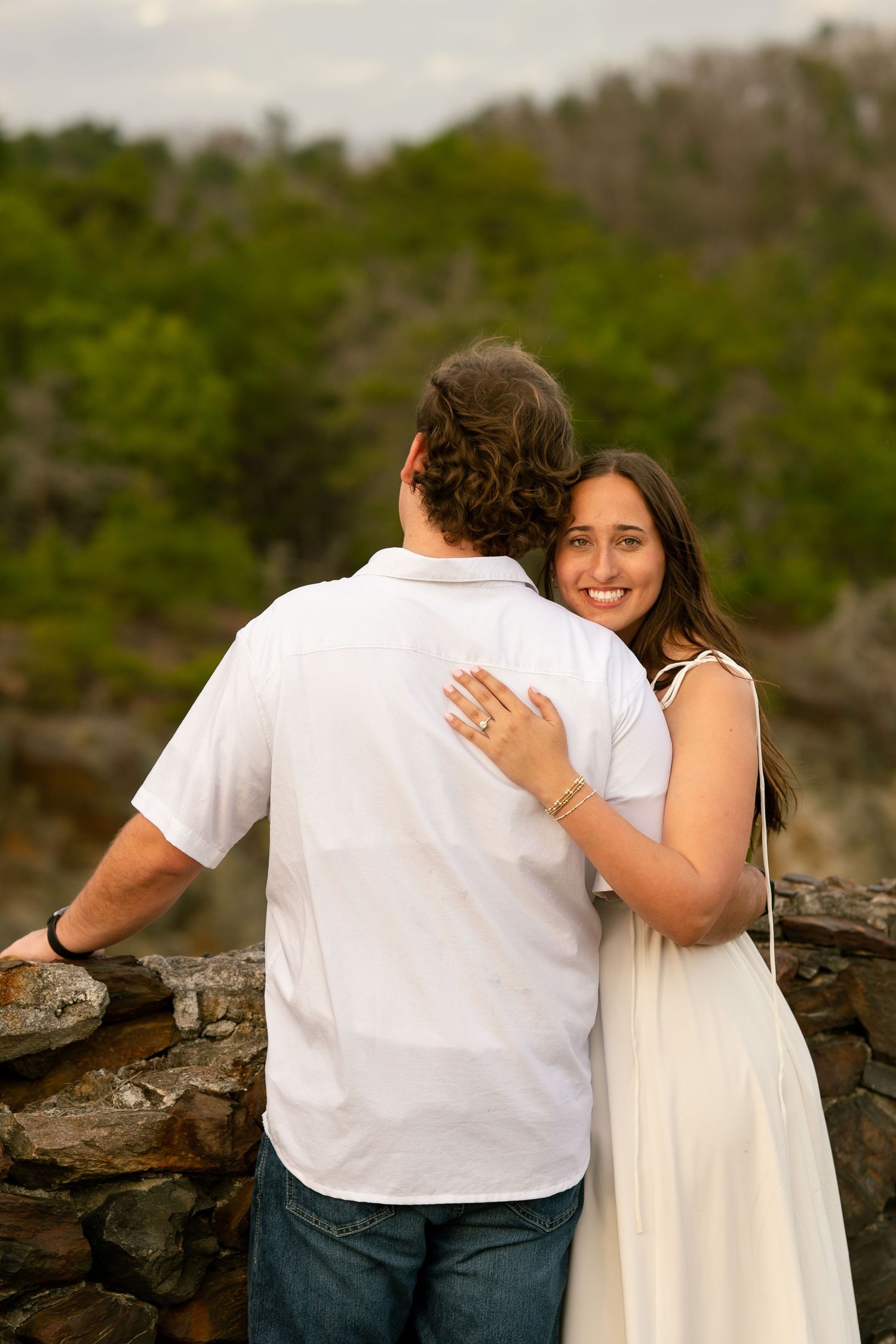 Smiling woman in white dress hugs man outdoors near stone wall with lush green trees in background.