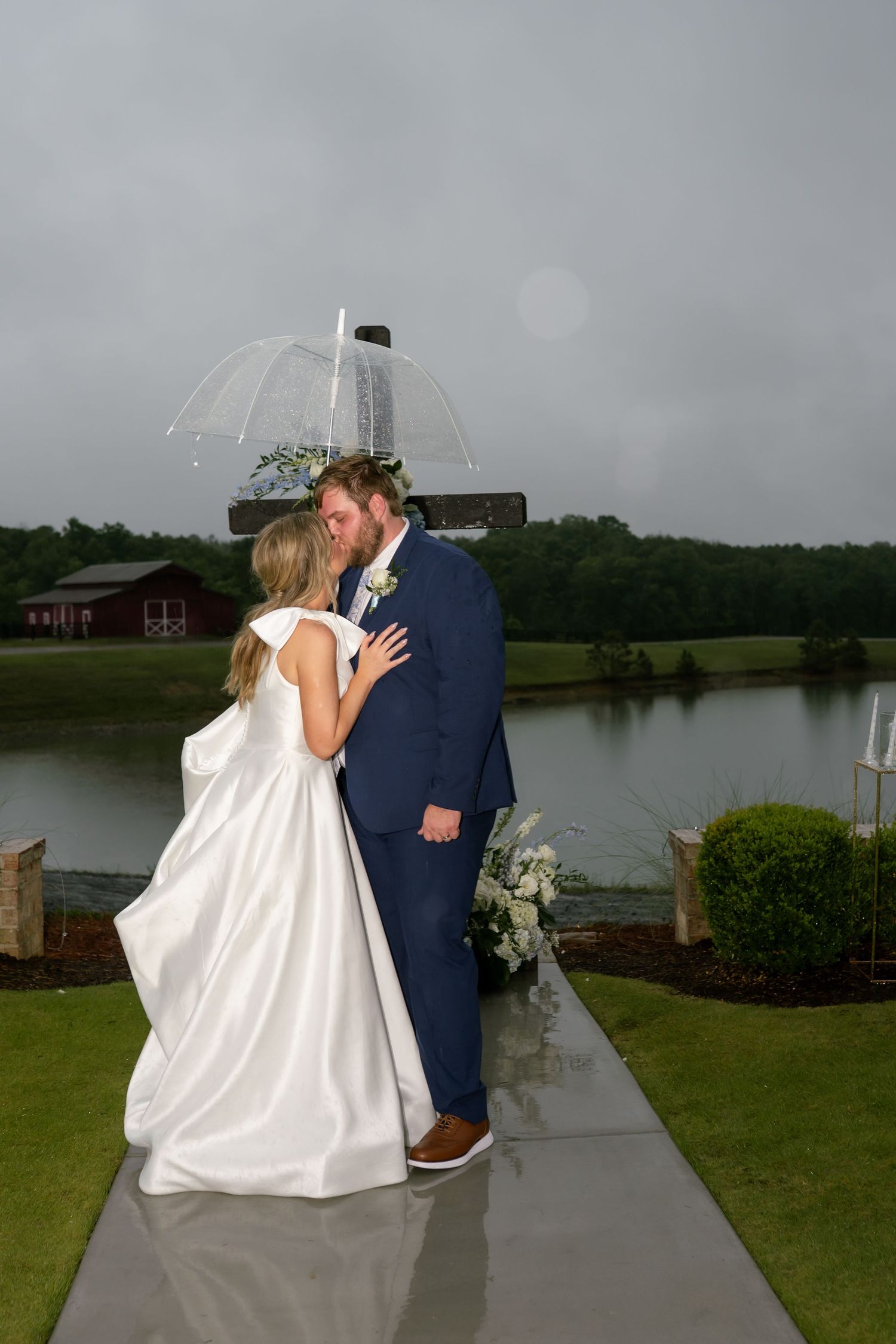 Bride and groom share a kiss under a clear umbrella on a rainy wedding day by a pond.