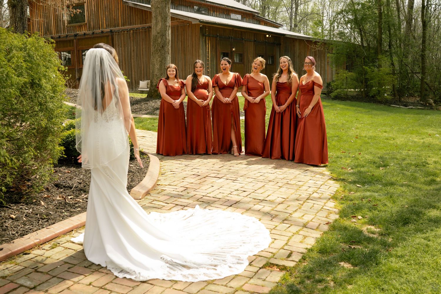 Bride in white gown and veil faces six bridesmaids in rust-red dresses on brick patio at rustic outdoor wedding venue.
