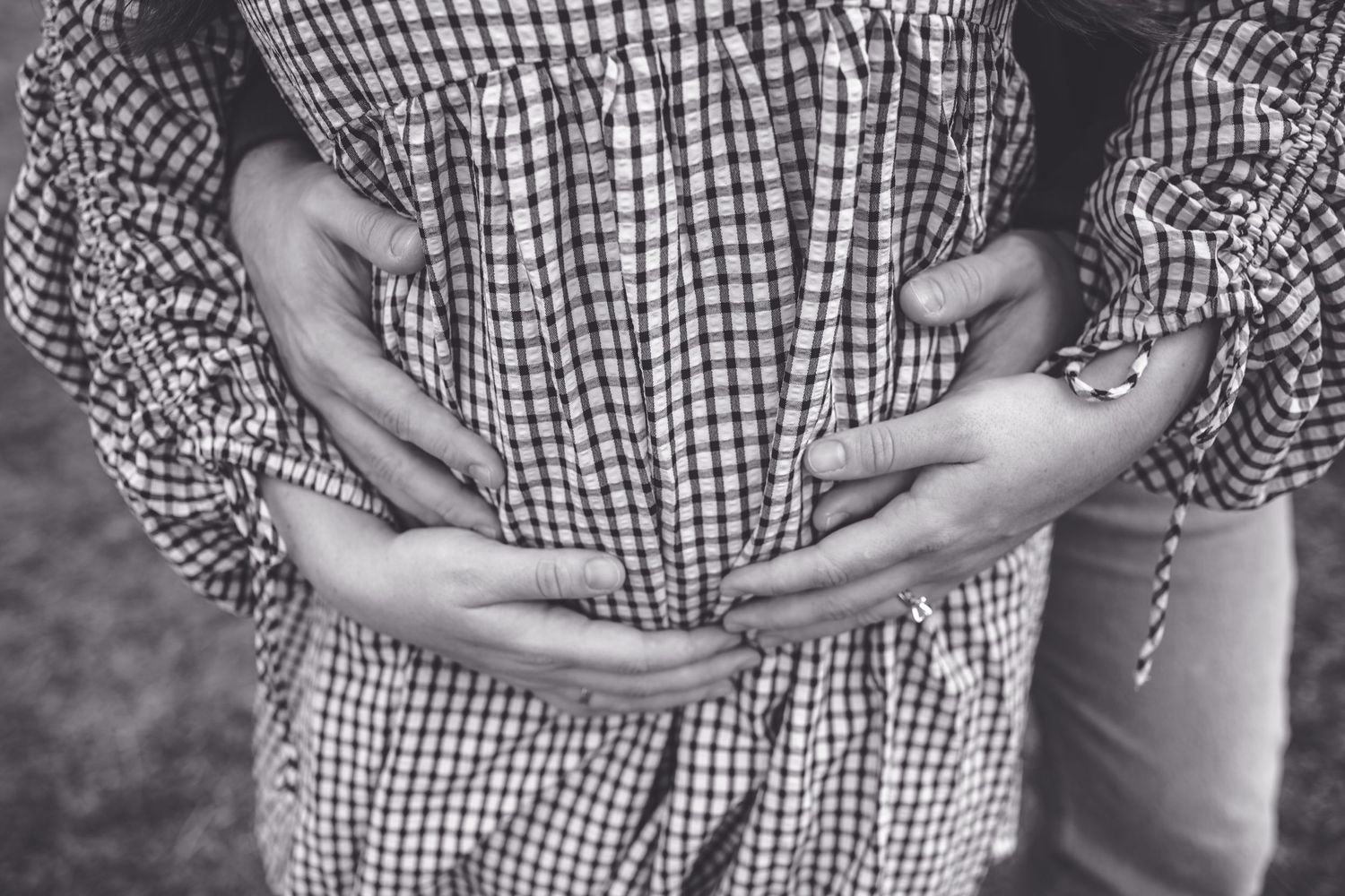 Black and white photo of hands cradling a pregnant belly wearing a gingham patterned dress.