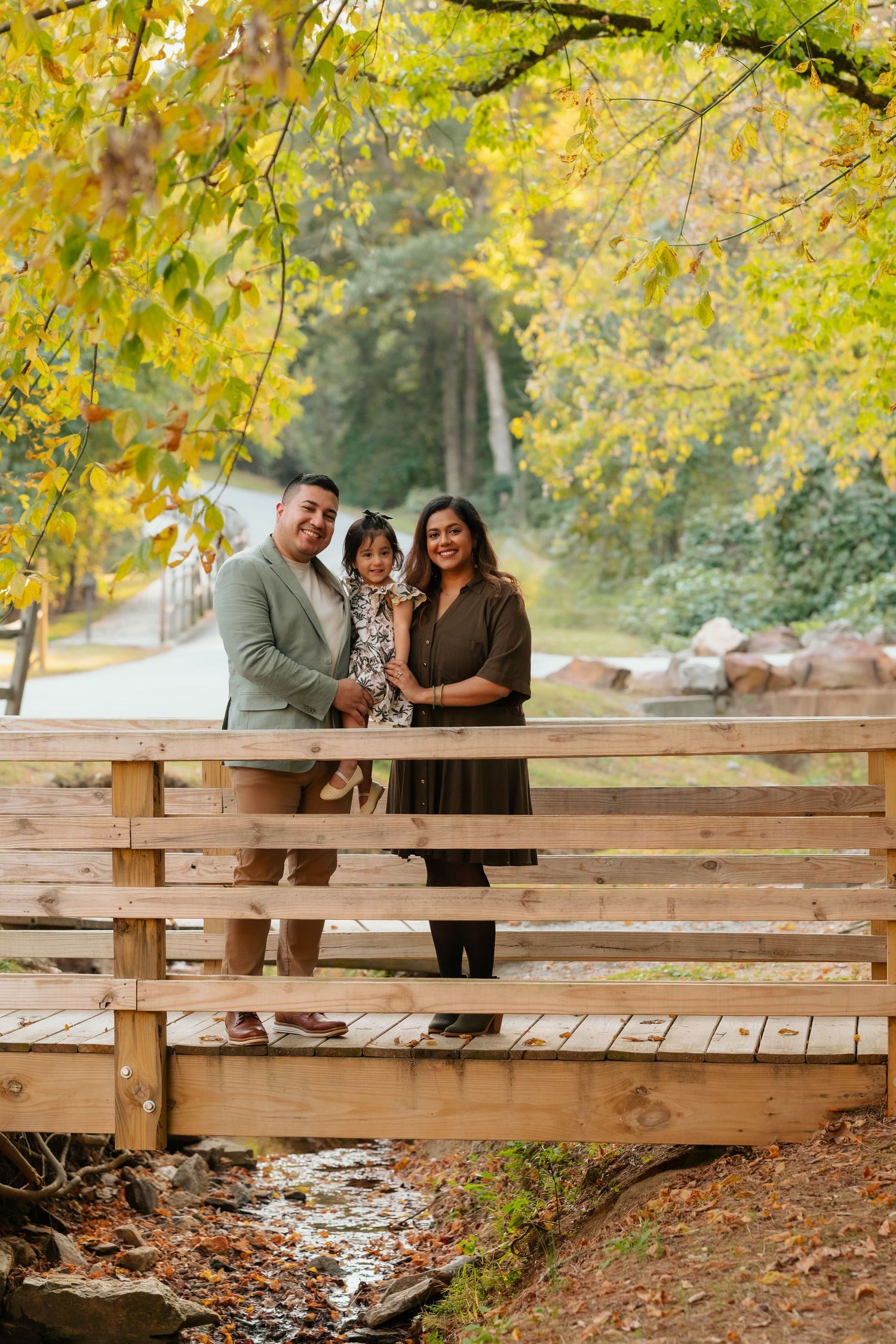 A family poses together on a wooden bridge surrounded by golden autumn leaves.