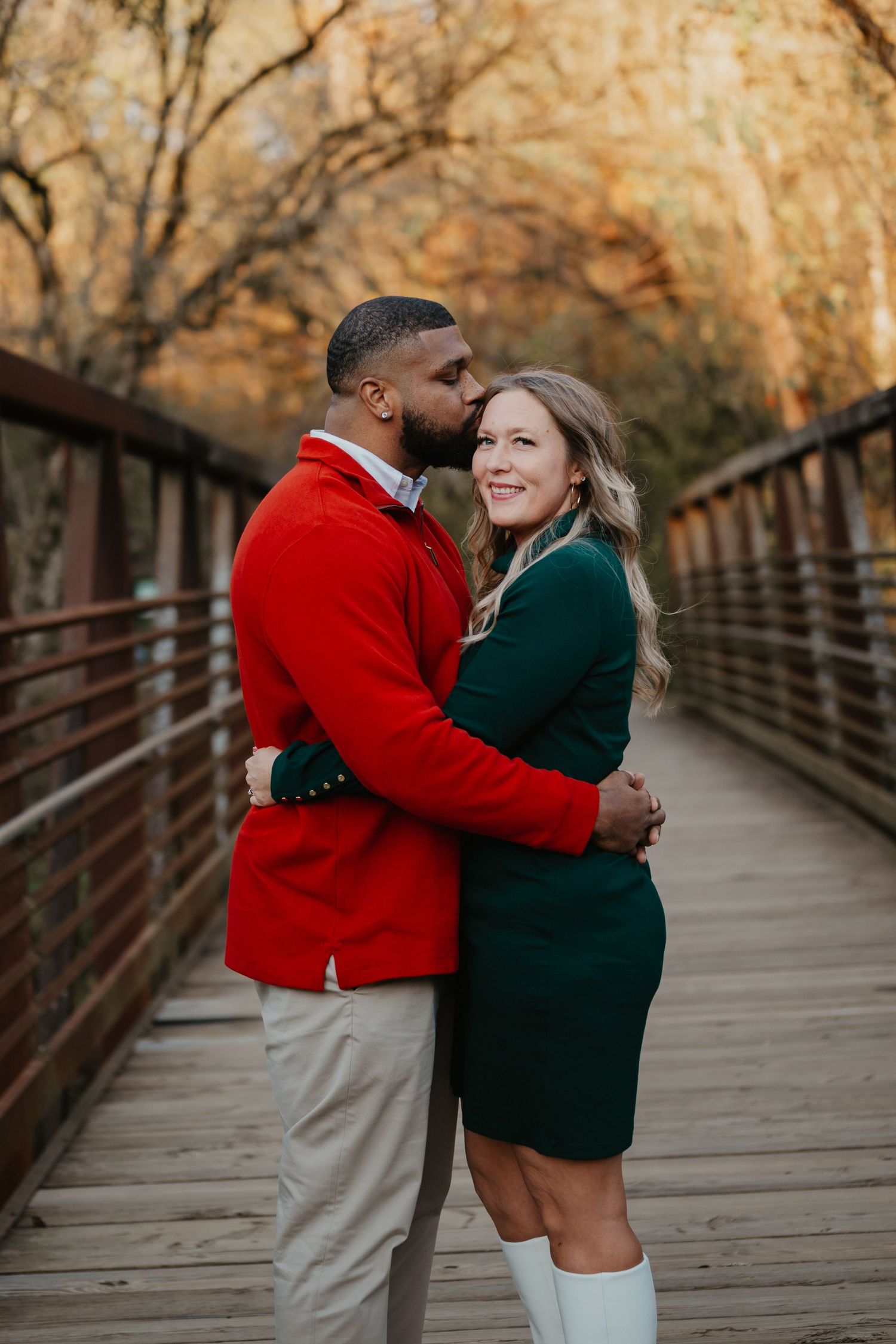 Couple embracing on wooden bridge wearing festive red and green outfits during autumn.
