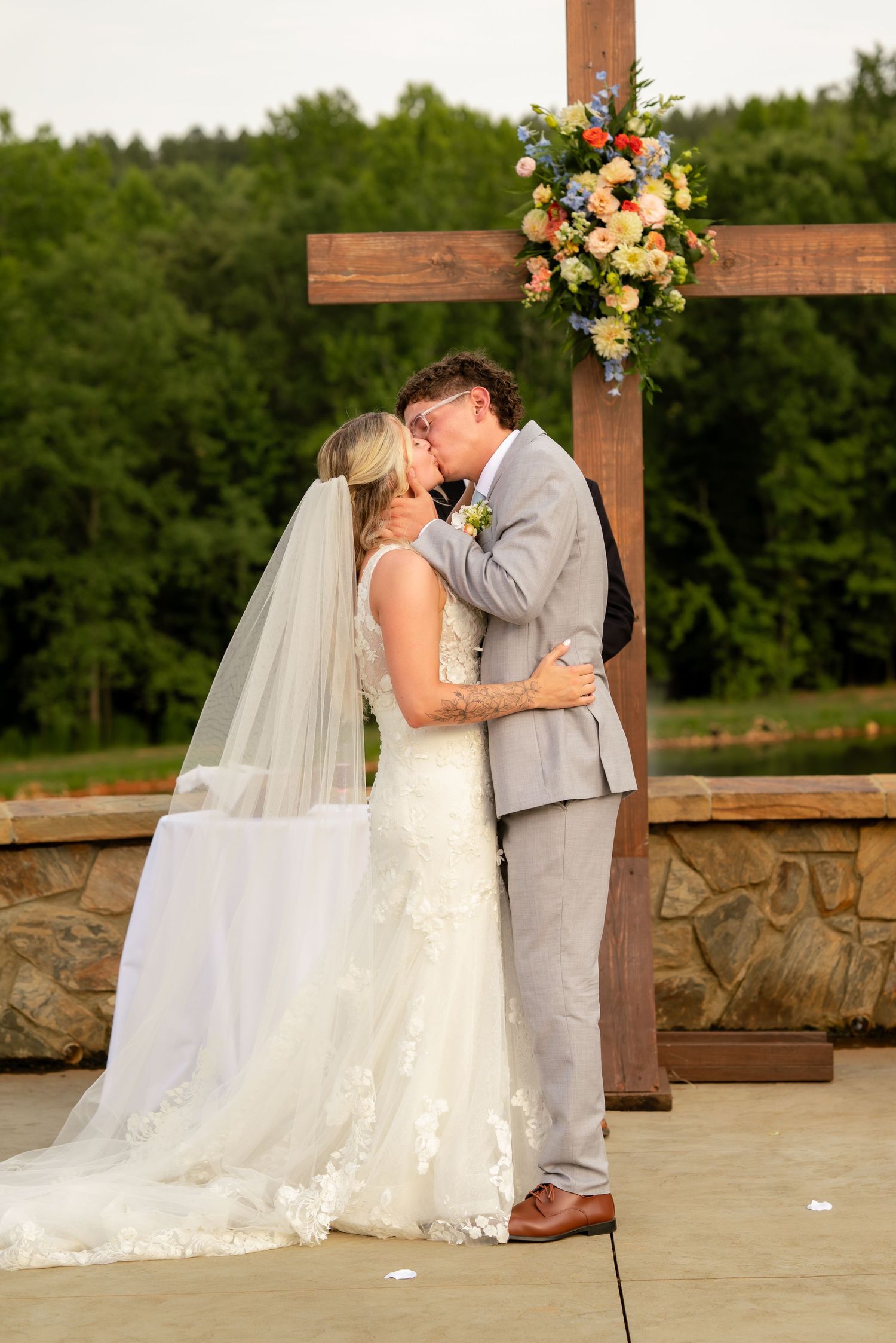 Bride and groom share first kiss at outdoor wedding ceremony beneath wooden cross adorned with colorful floral arrangement.