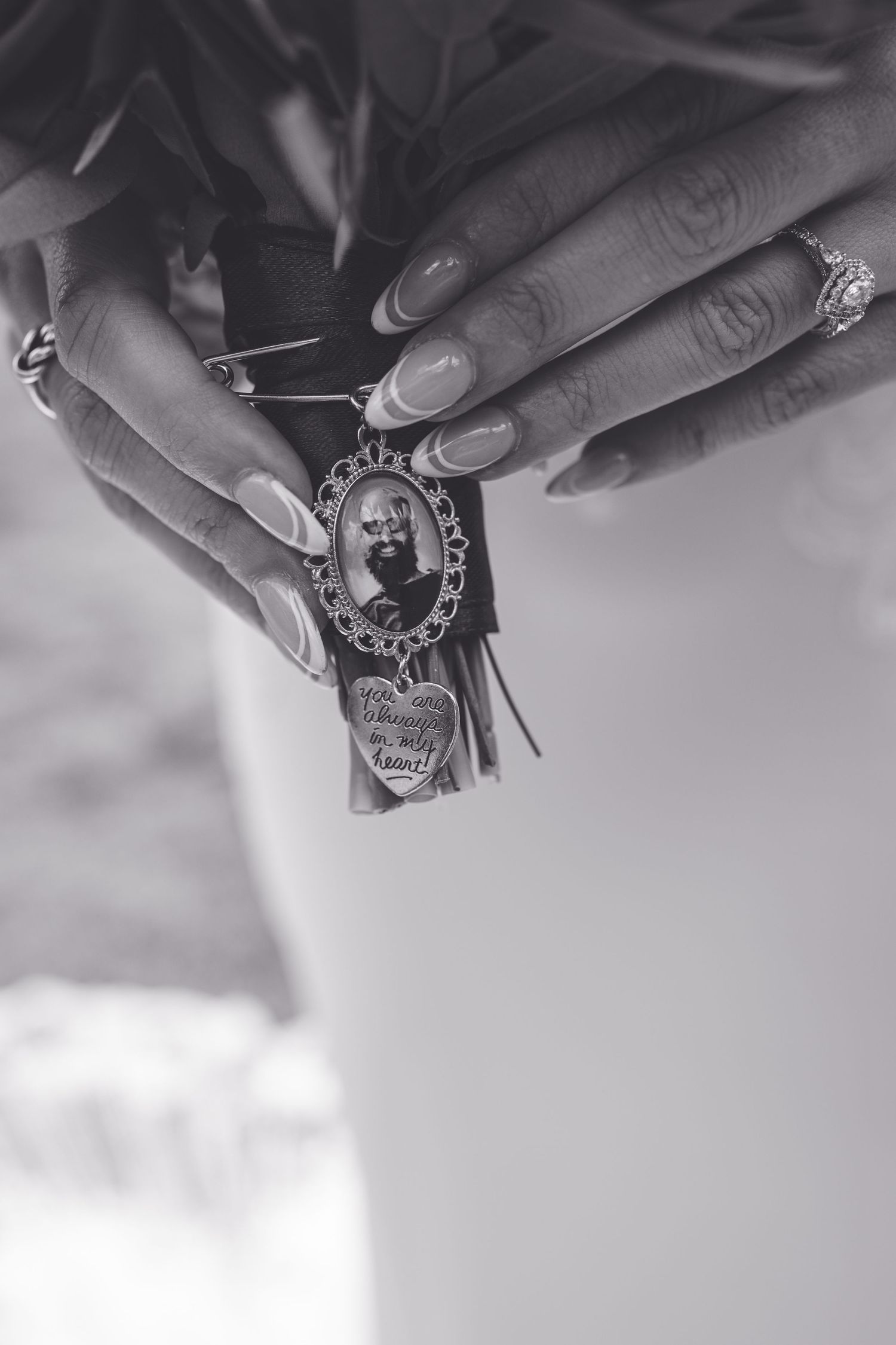 Black and white close-up of bride holding bouquet with a memorial photo charm pendant in manicured hands.