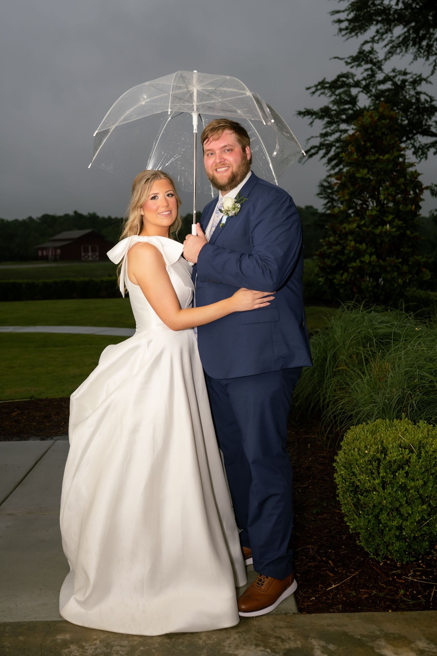 Bride and groom posing under clear umbrella in rain, bride in white gown, groom in navy suit with boutonniere.