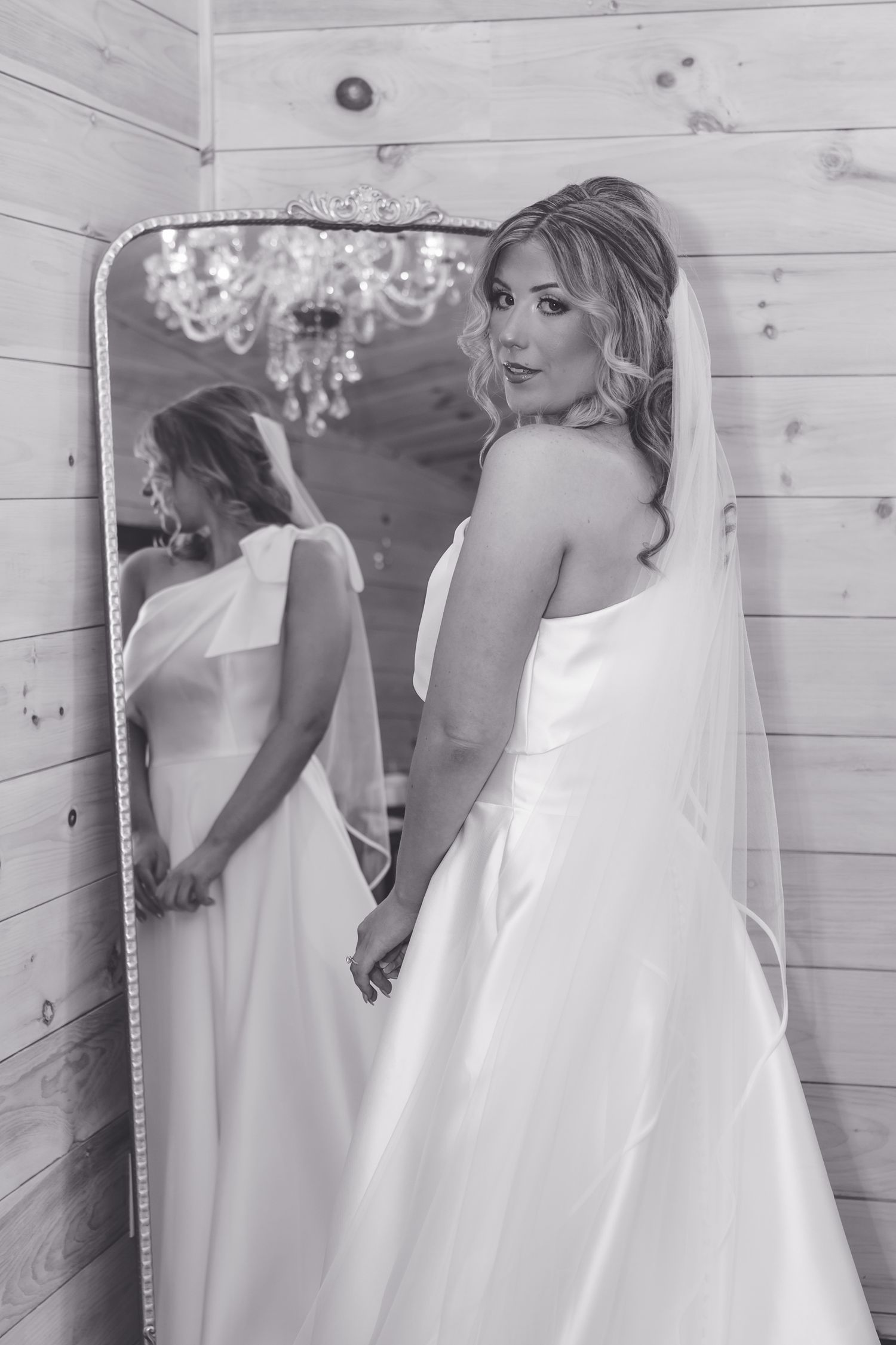 Bride in white gown and veil poses beside ornate mirror in black and white wedding photo.
