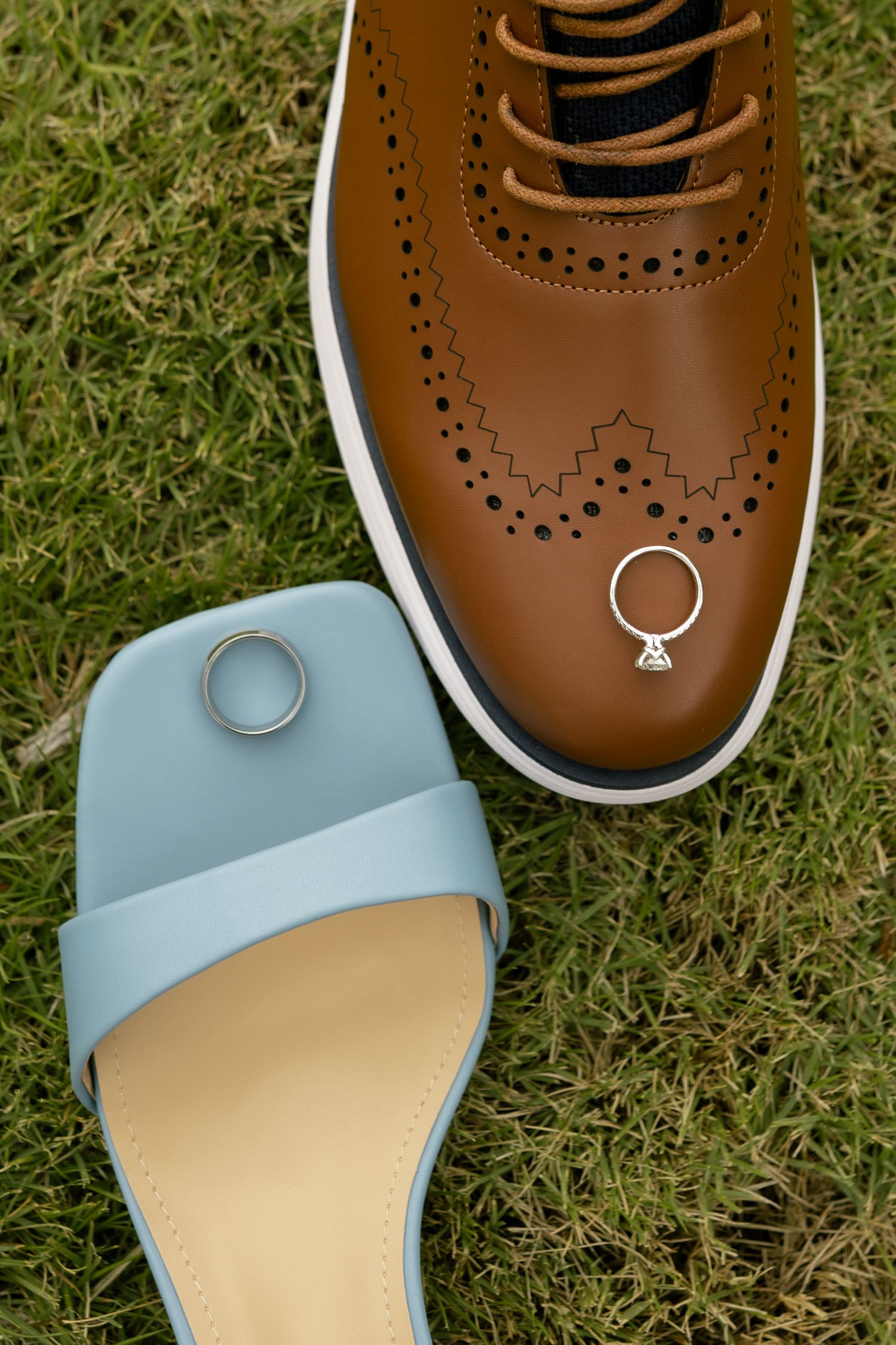 Brown brogue dress shoe and blue sandal on grass with wedding rings resting on each shoe.