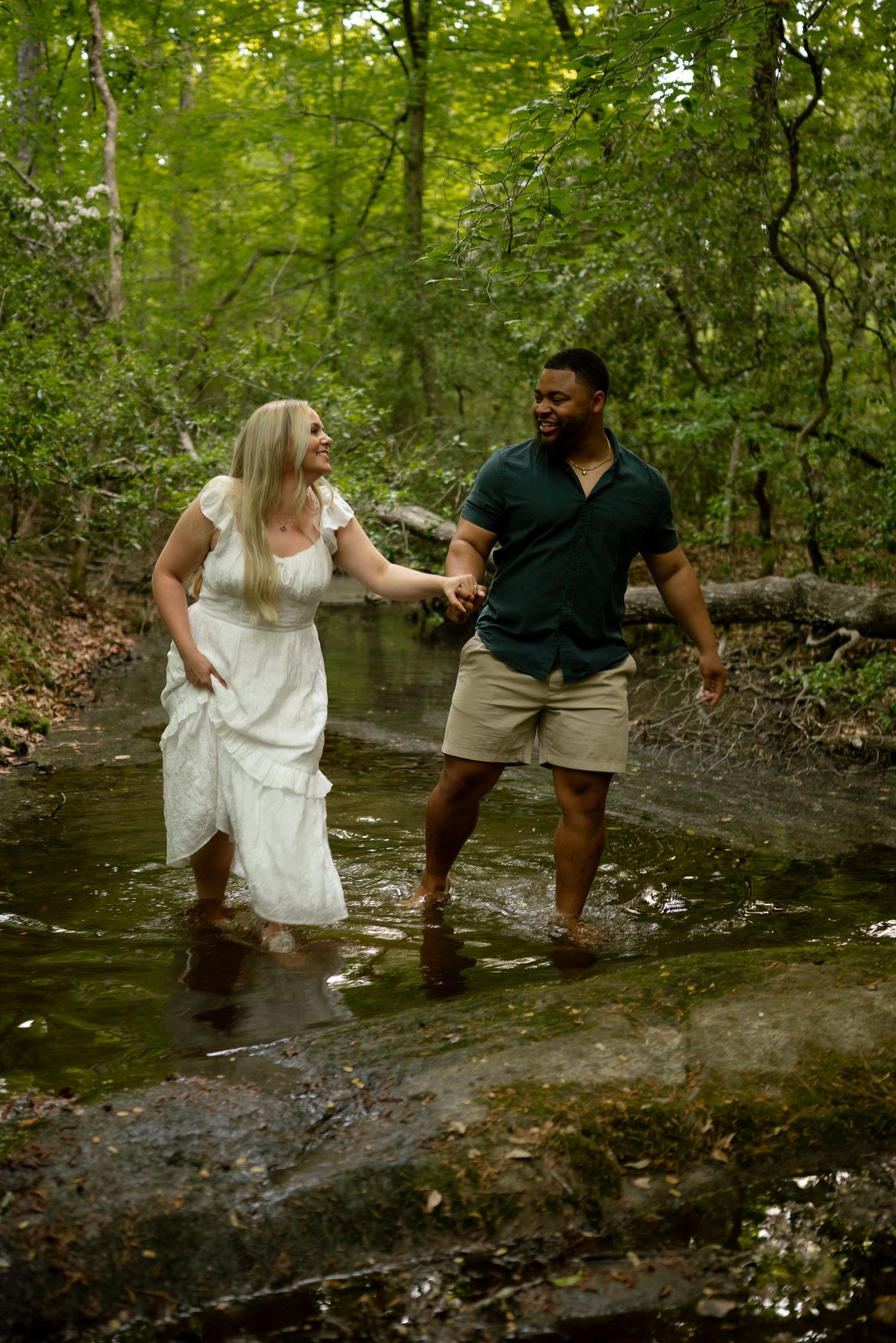 Couple holding hands wading through a forest creek, woman in white dress, man in green shirt and khaki shorts.