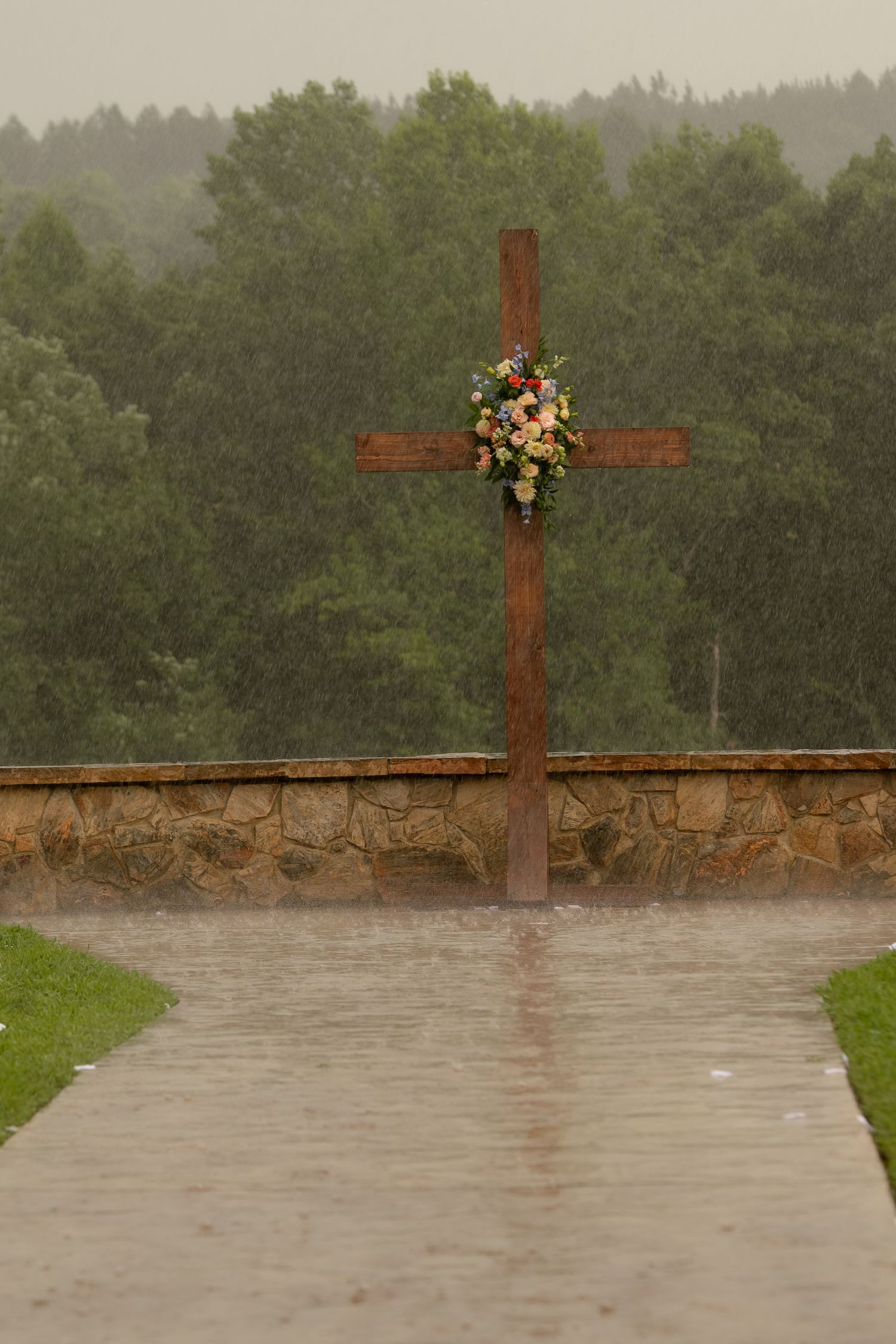 Wooden cross with floral wreath stands in heavy rain, stone wall and green trees in background.