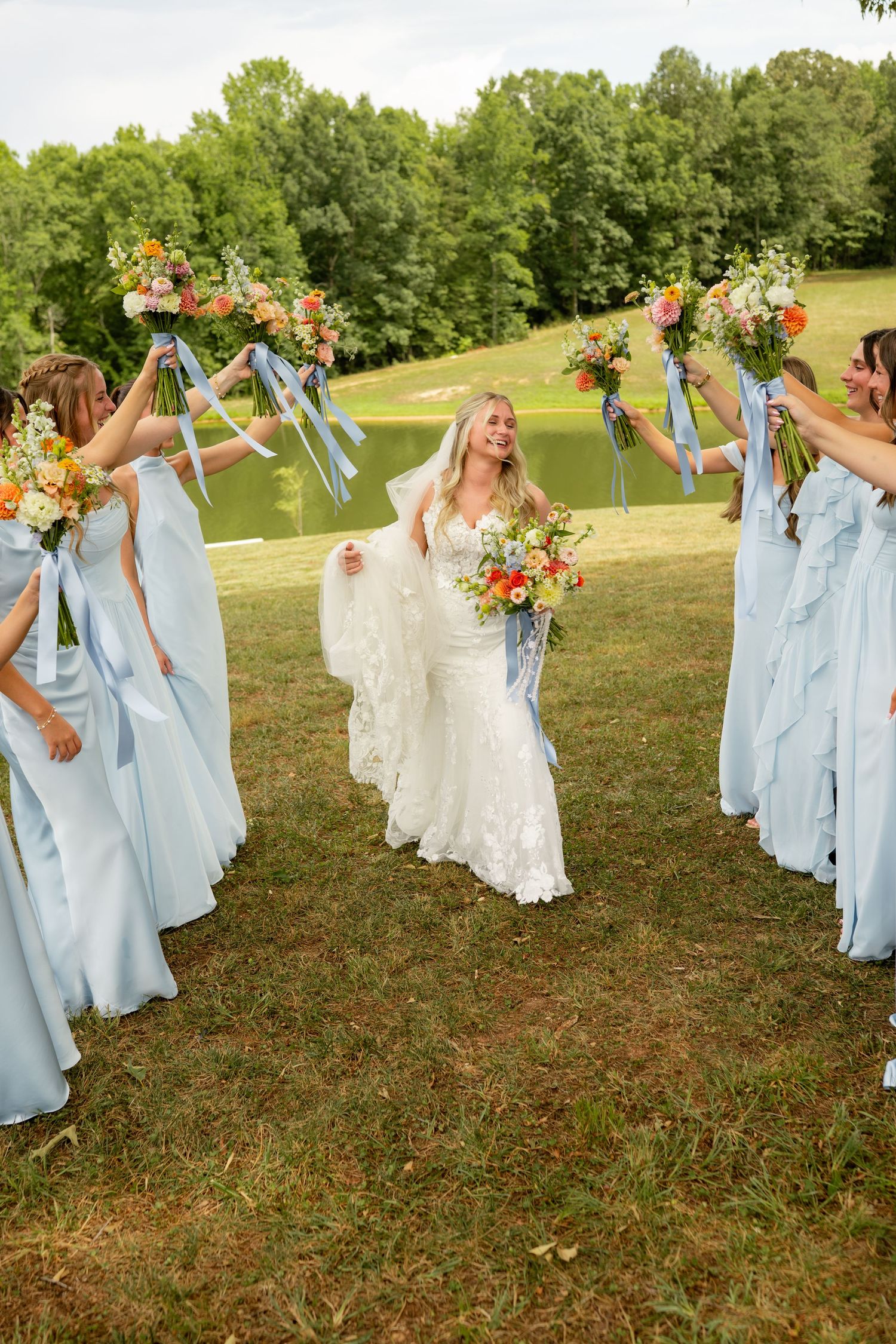 Bride in lace gown walks through tunnel of bridesmaids in blue dresses raising colorful bouquets at outdoor wedding.