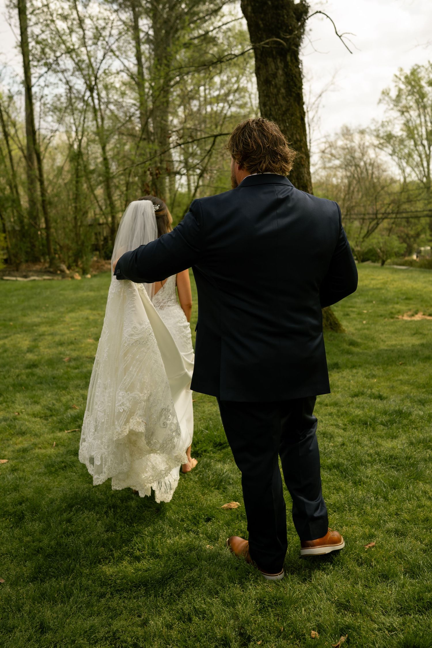 Bride and groom walking away hand in hand across green grass, her lace veil trailing behind.