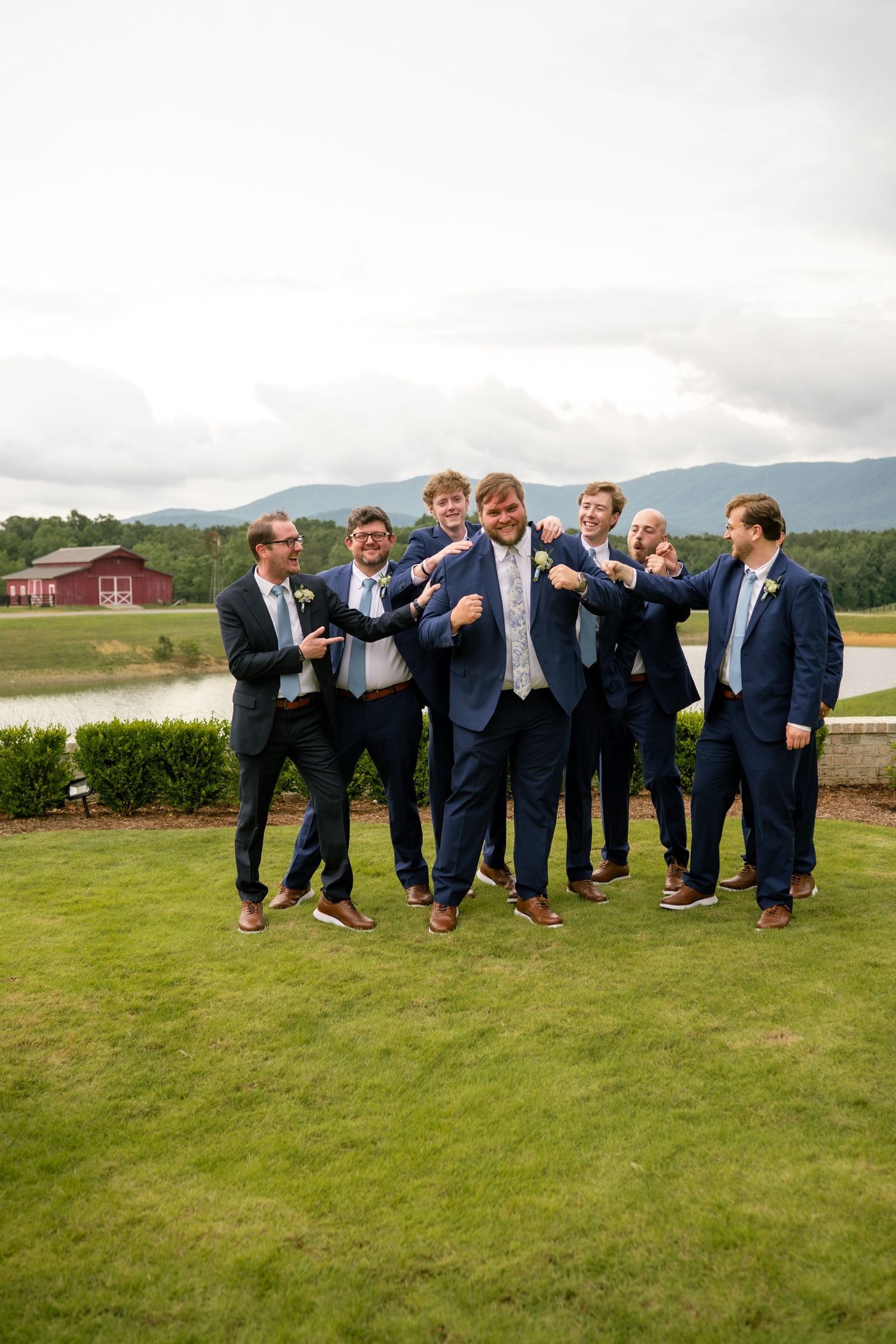 Groomsmen in navy suits playfully surrounding groom outdoors with scenic mountain landscape and red barn in background.
