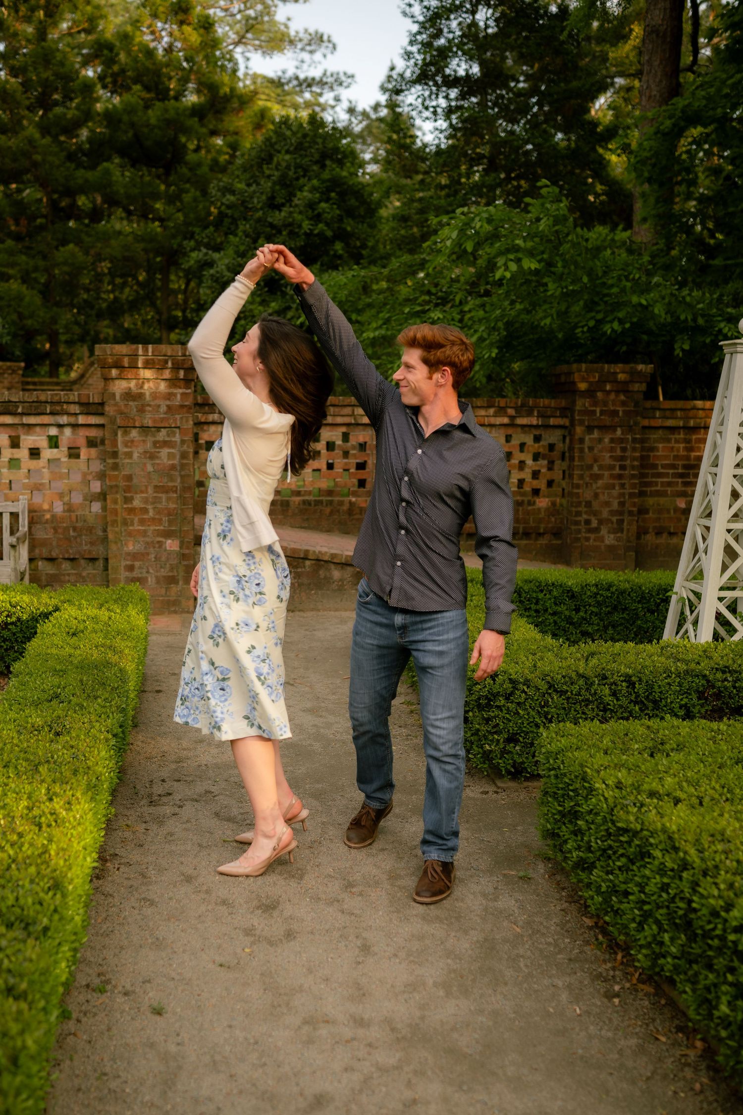 Couple dancing together in a lush garden pathway, man spinning woman in floral dress near brick wall.